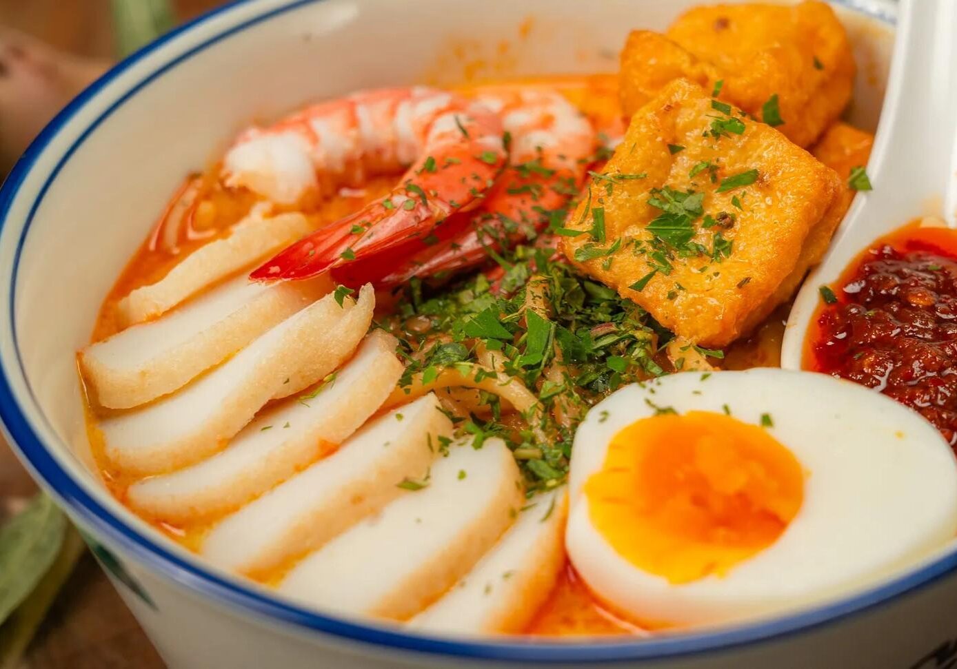 This image features a vibrant bowl of Laksa, a spicy Southeast Asian noodle soup topped with fresh prawns, sliced fish cakes, and golden-brown fried tofu puffs. The dish is garnished with finely chopped herbs and served with a soft-boiled egg and a side of dark red sambal chili for an extra kick of flavor.