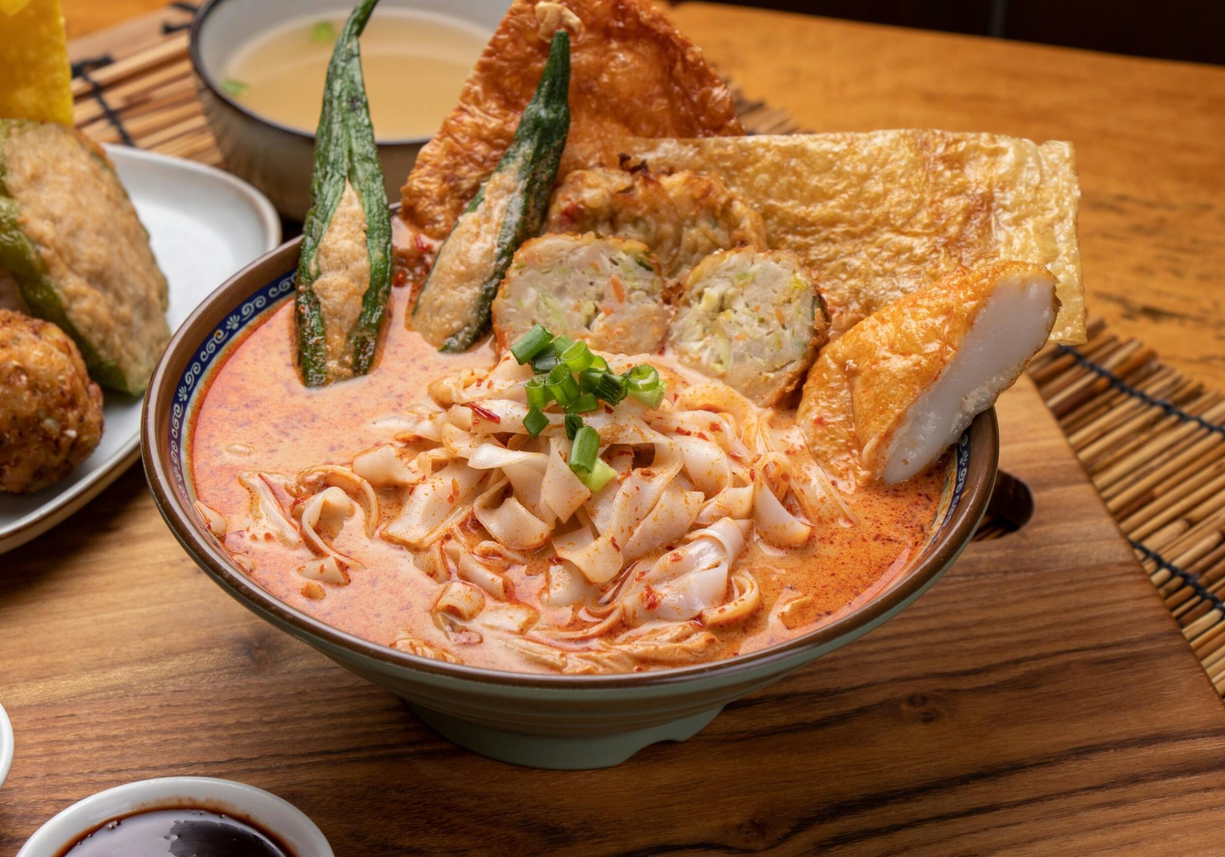 A bowl of flat rice noodles is served in a creamy, orange-tinted spicy broth and topped with various fried items, including stuffed okra and fish cakes. The dish is presented on a wooden table alongside small side plates of additional fried appetizers and dipping sauces.