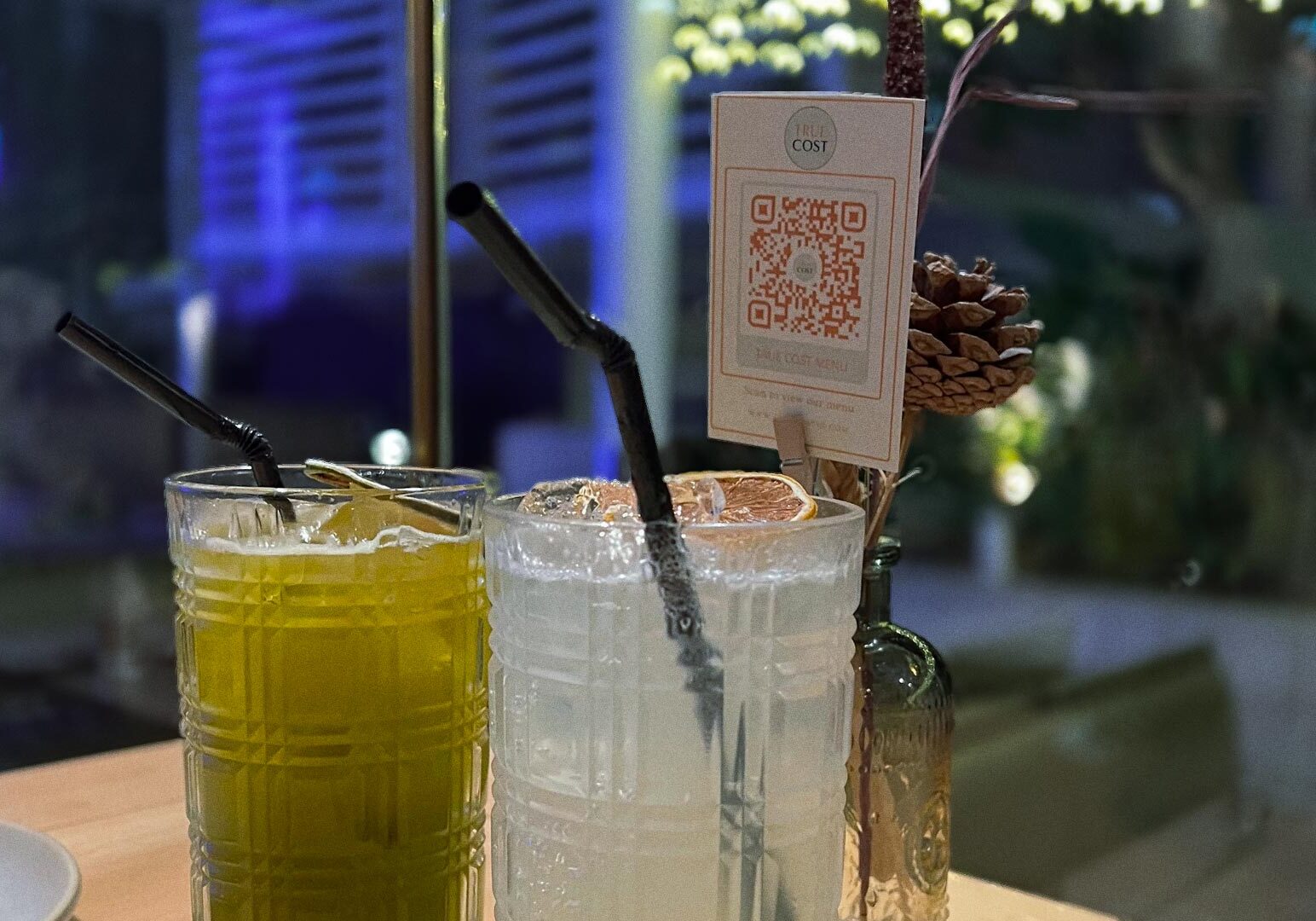 Two colorful iced drinks at True Cost Suntec City beside a decorative QR code card in a dim, cozy setting.