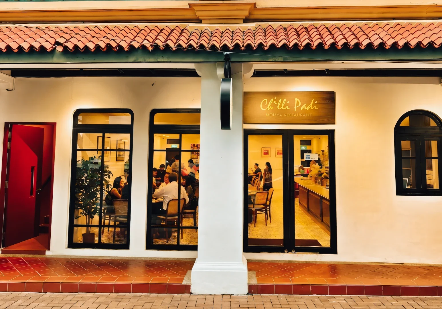 Chilli Padi Nonya Restaurant exterior showcasing white shophouse façade, warm window lighting, tiled walkway, and diners visible through the glass windows.