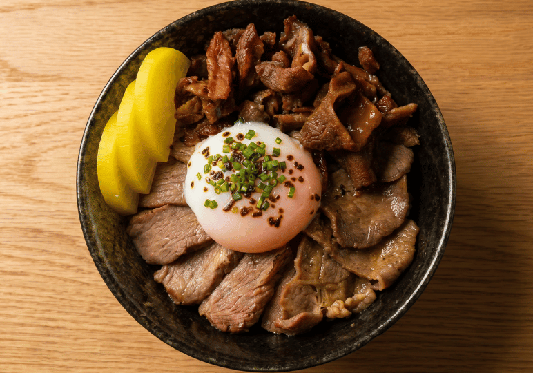 A close-up, top-down view of a Japanese rice bowl topped with sliced beef, braised meat, bright yellow pickled radish, and a soft-boiled egg garnished with herbs and seasoning.