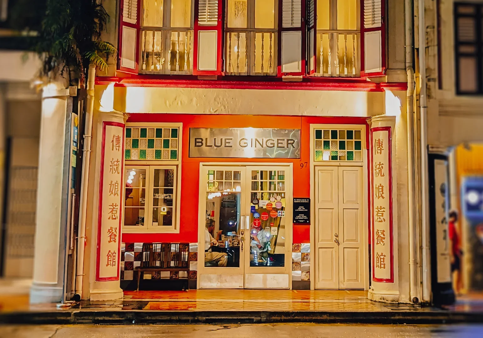 Front exterior of Blue Ginger restaurant featuring red-and-white shophouse architecture, traditional Chinese vertical signage panels, warm interior lighting, and a glass entrance.