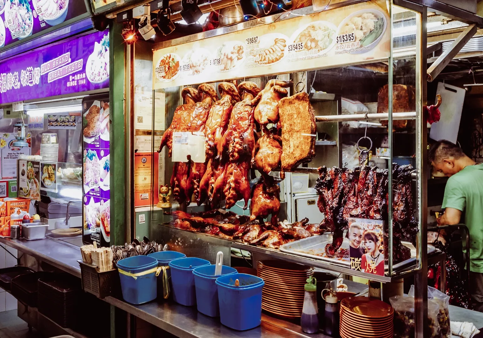 Eye‑level wide shot of a bustling Singapore hawker stall displaying roasted meats hanging in a glass case, with a vendor preparing charred meats for traditional street food dining.