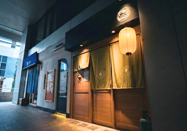 8-orchard-plaza-stalls The discreet, traditional wooden entrance of a Japanese restaurant in the Orchard Plaza hallway, featuring green noren curtains and a glowing paper lantern.