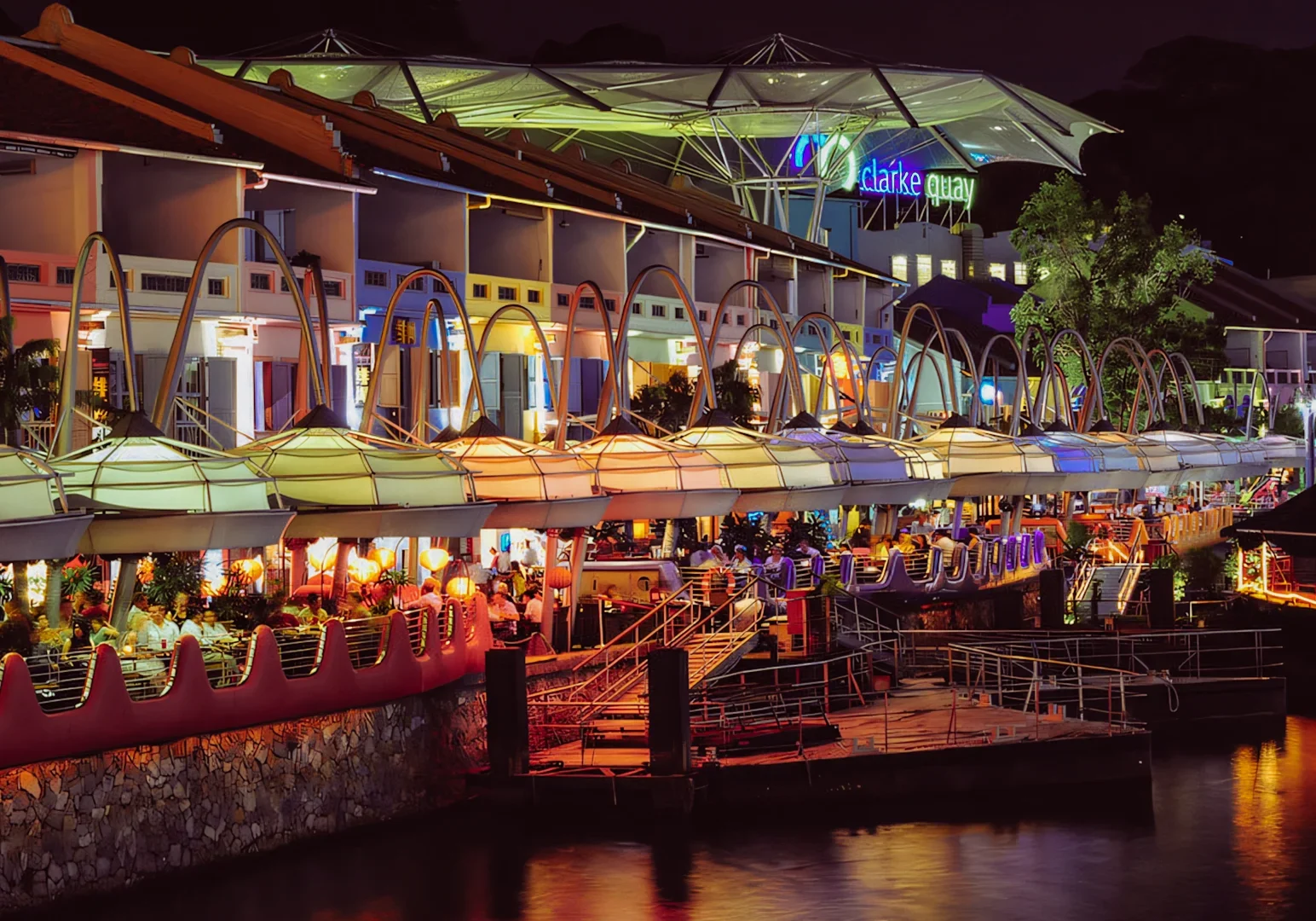Wide‑angle nighttime shot of Clarke Quay in Singapore, showing lively waterfront restaurants with illuminated facades, crowds dining outdoors, and reflections along the Singapore River.