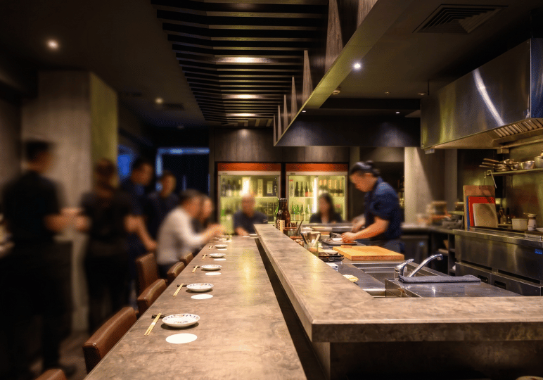 Interior of a Japanese restaurant featuring a long dining counter where patrons are seated while chefs prepare food in the open background kitchen.