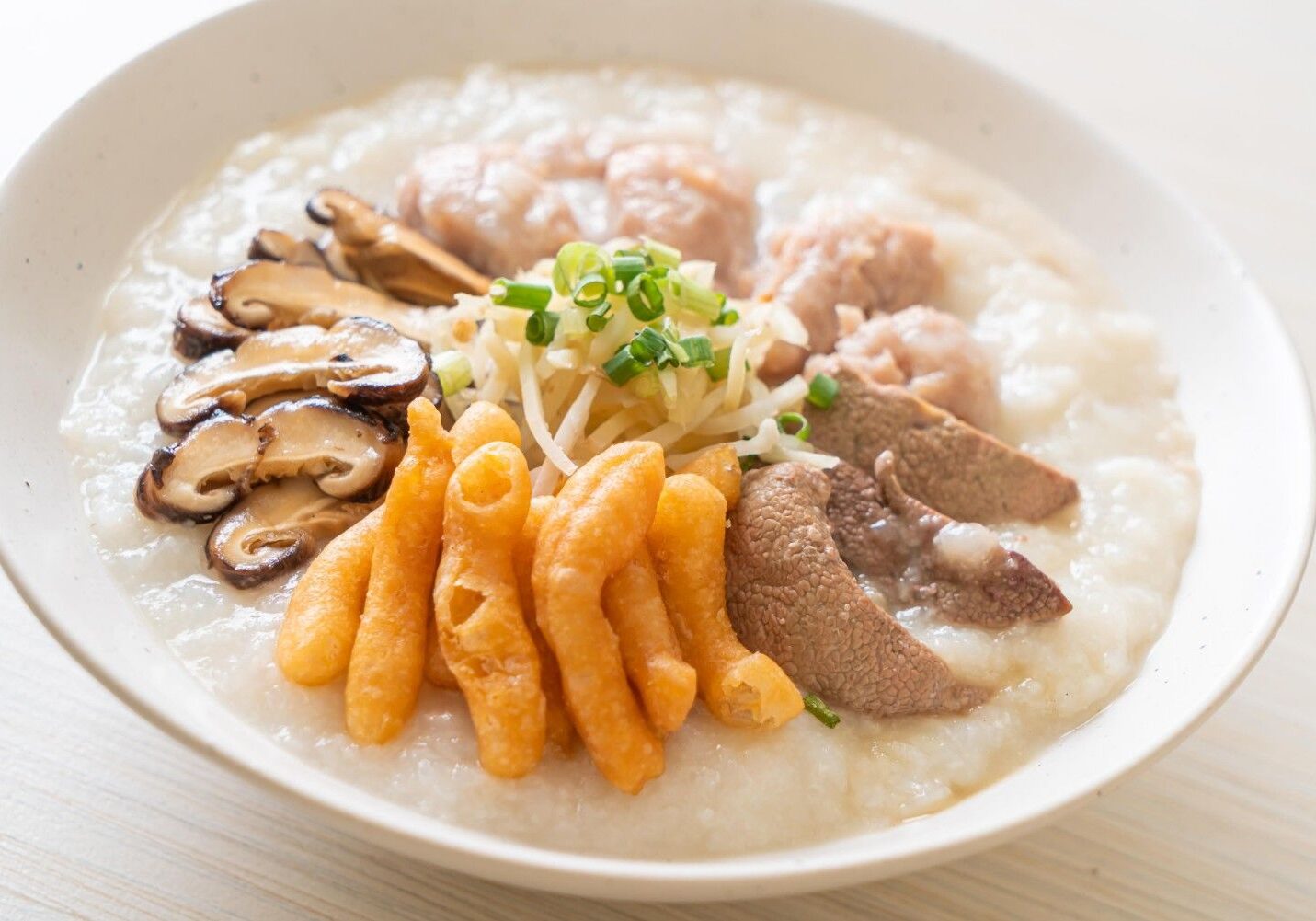 This appetizing bowl of rice congee features a hearty assortment of toppings, including pork meatballs, sliced liver, savory shiitake mushrooms, and crispy fried dough sticks. The porridge is finished with a fresh garnish of julienned ginger and chopped scallions, presented simply in a white ceramic bowl.