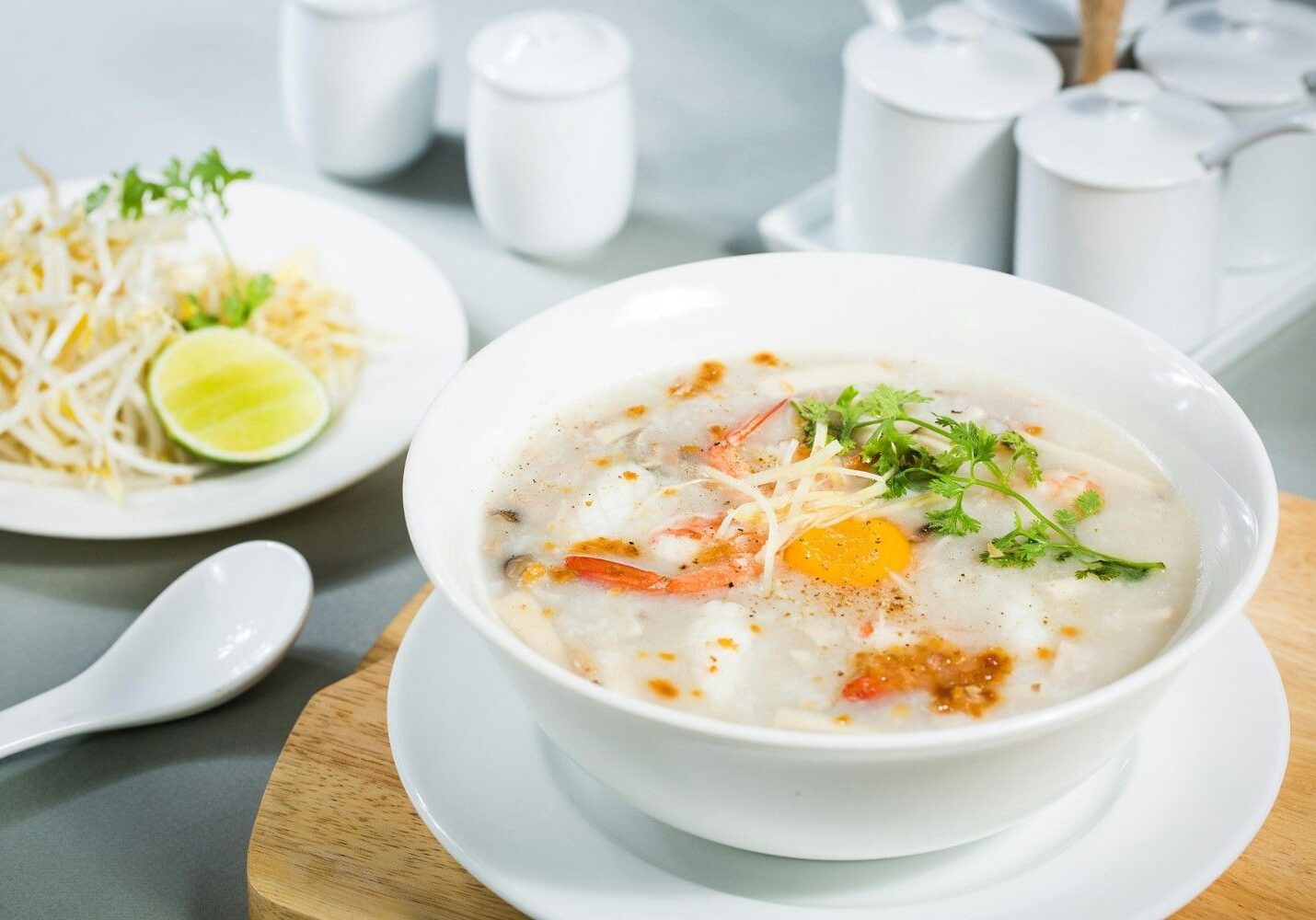 This image features a bowl of savory rice porridge containing shrimp and a bright egg yolk, garnished with ginger strips, fried garlic, and fresh cilantro. The dish is served on a wooden board alongside a spoon and a separate plate of bean sprouts and lime, with condiment jars visible in the background.