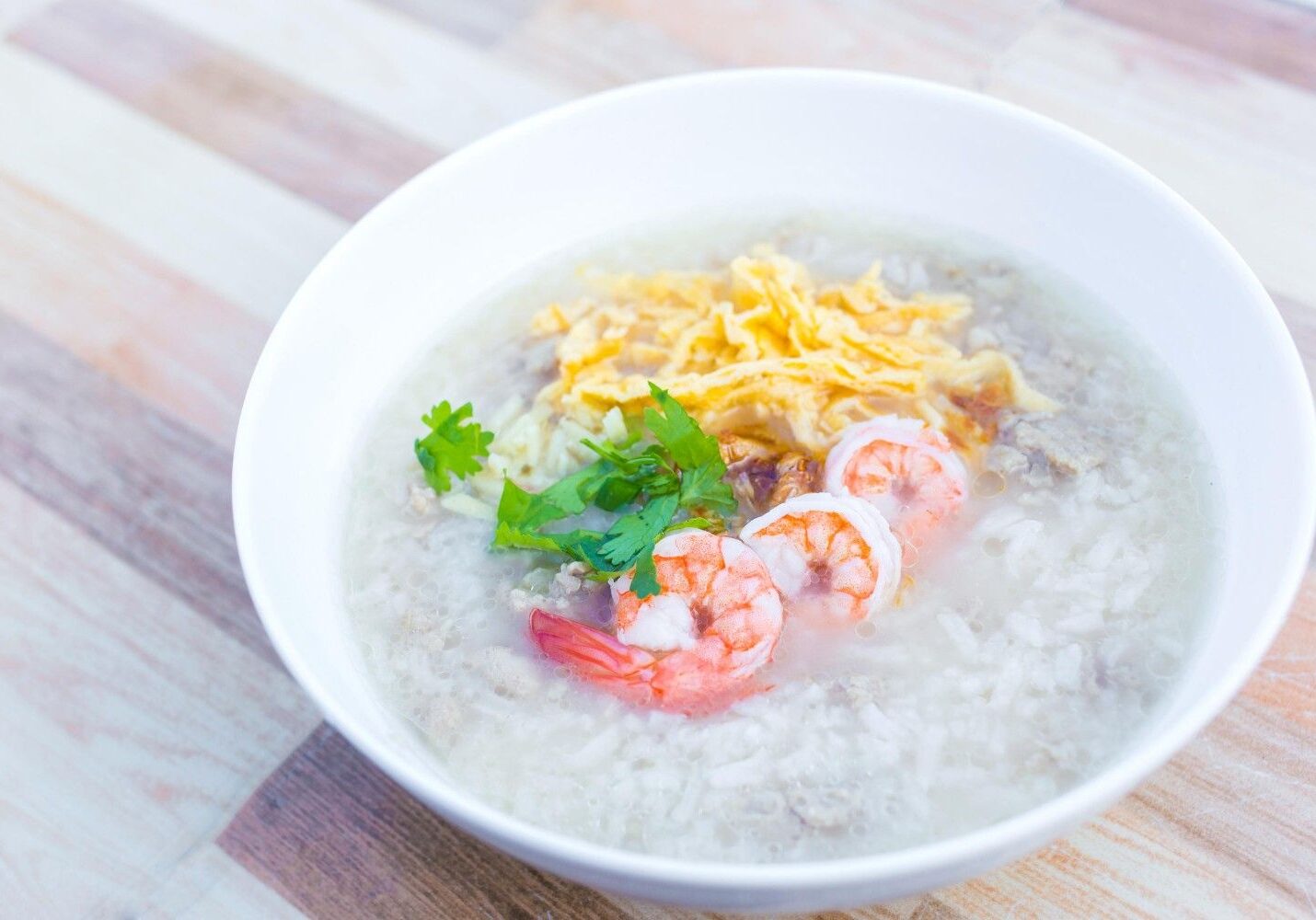 This image features a white bowl filled with clear rice porridge that includes ground pork and three cooked shrimp. The savory dish is topped with shredded yellow omelet, crispy fried garlic, and fresh cilantro leaves, set against a light striped wooden background.