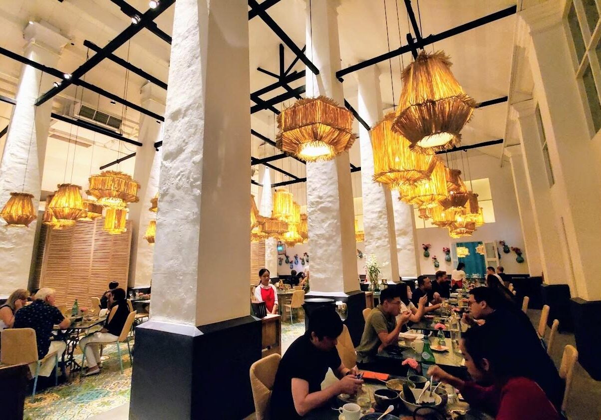 This expansive restaurant interior features soaring white textured columns and an array of warm, straw-like pendant lanterns hanging from a dark industrial ceiling. Patrons are seated at various tables across a patterned tile floor, enjoying their meals in a bright, high-ceilinged space that blends rustic and modern design elements.