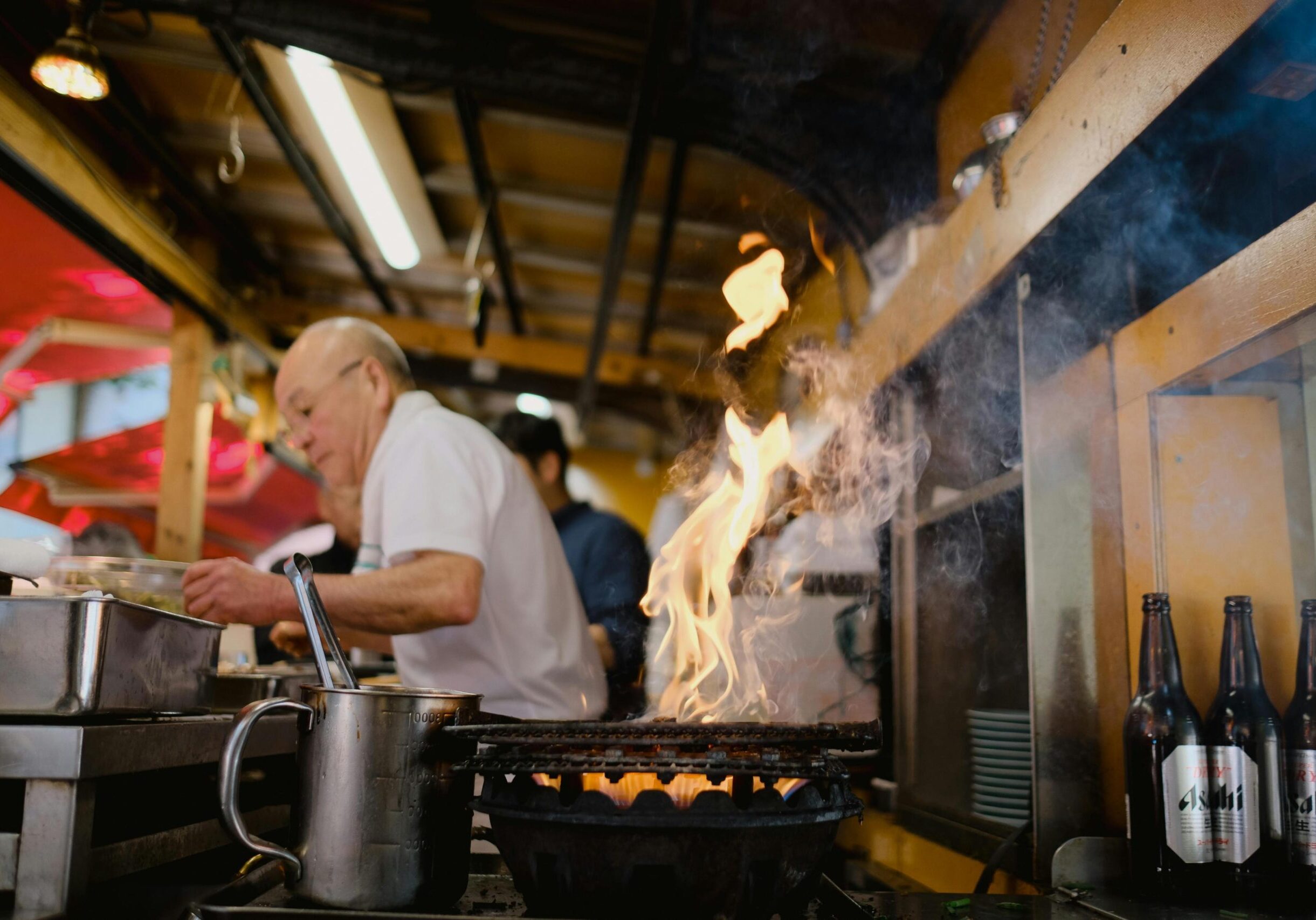 Bright orange flames and swirling smoke rise dramatically from a grill, taking center stage in this bustling kitchen scene. In the background, a chef diligently prepares food near a row of Asahi beer bottles, creating an atmosphere typical of a lively izakaya or street food stall.