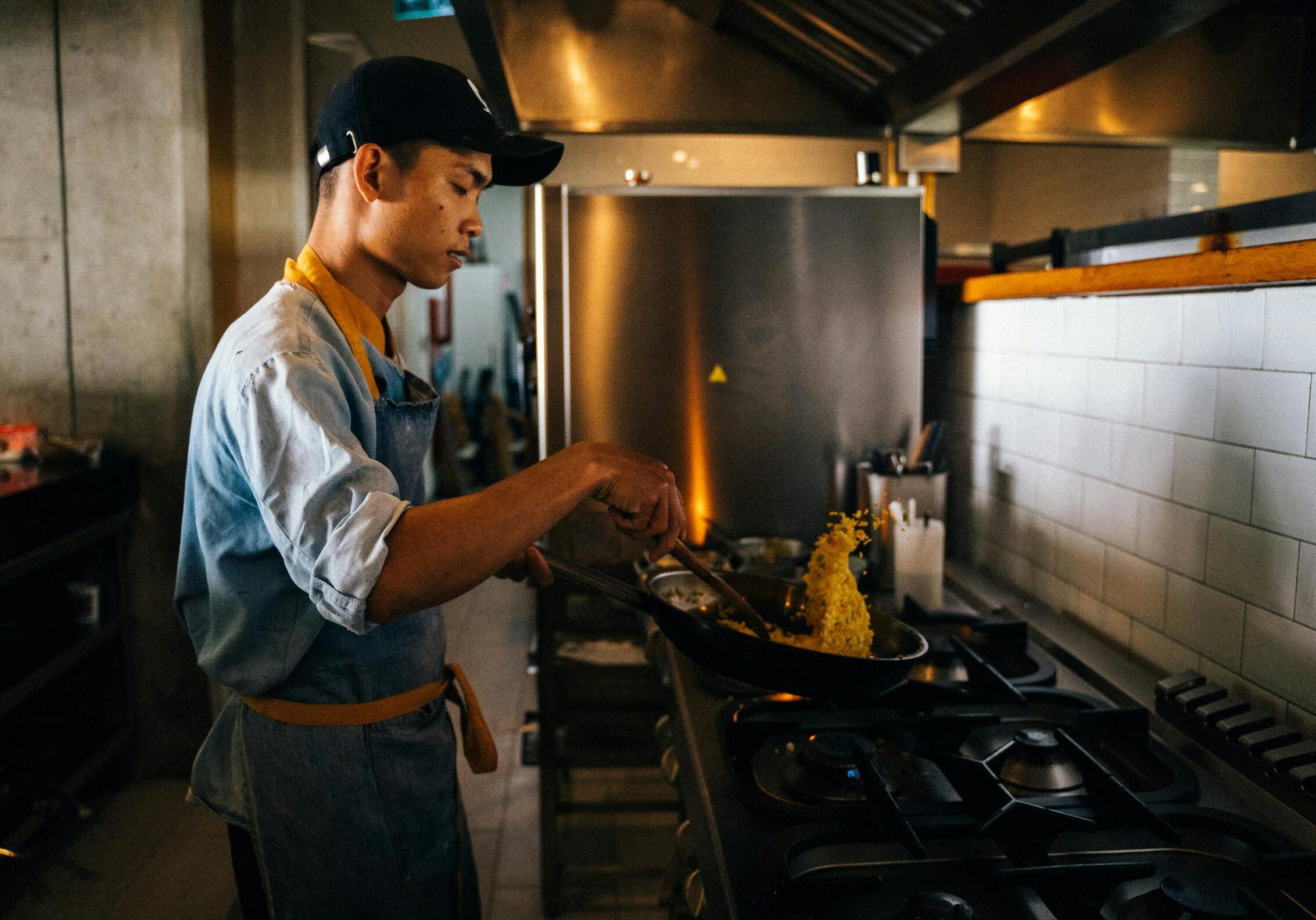 Captured mid-action, a focused chef wearing a black cap and blue apron skillfully tosses yellow ingredients in a frying pan atop a commercial gas stove. The scene takes place in a professional kitchen characterized by stainless steel appliances and white subway tiles, highlighting the cook's concentration.