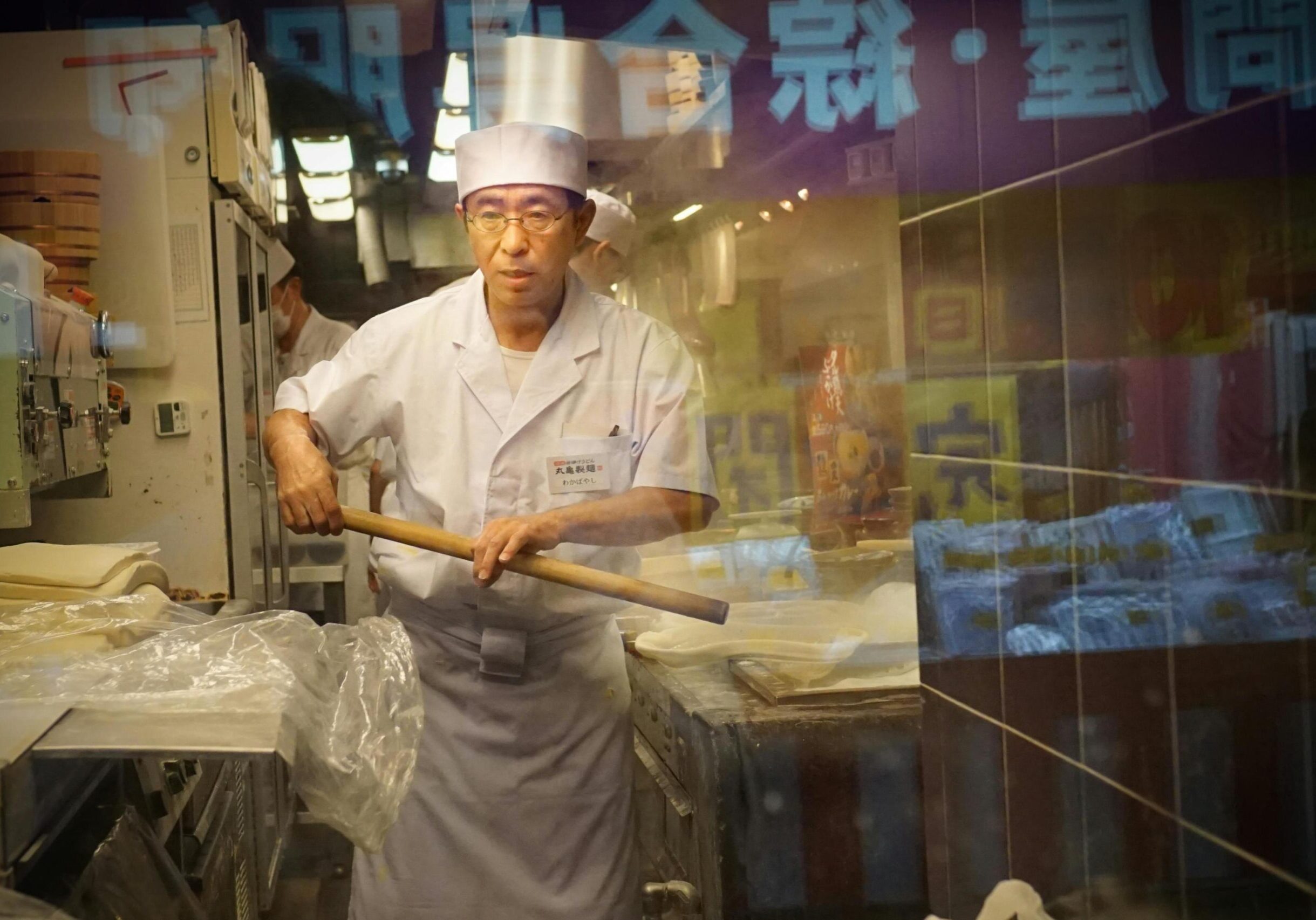 A focused chef wearing a white uniform and glasses stands behind a glass partition, holding a long wooden rolling pin typically used for noodle making. The windowpane reflects bright purple street signs with foreign characters, creating a visual layer that merges the busy exterior with the quiet concentration of the kitchen.