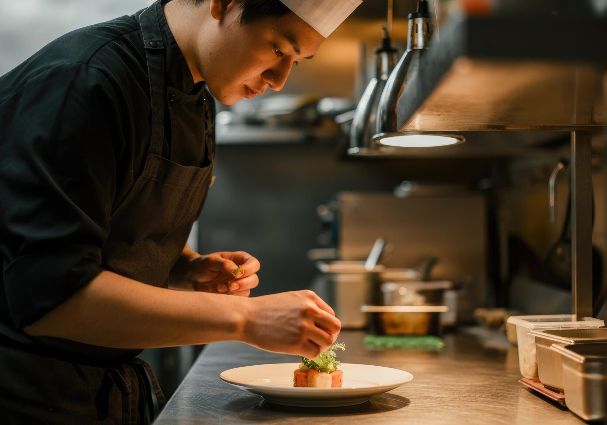 This close-up shot portrays a concentrated chef in a black uniform meticulously plating a delicate dish within a professional kitchen setting. Warm lighting from above highlights his hands as he carefully places a final green garnish, underscoring the precision and attention to detail in his craft.