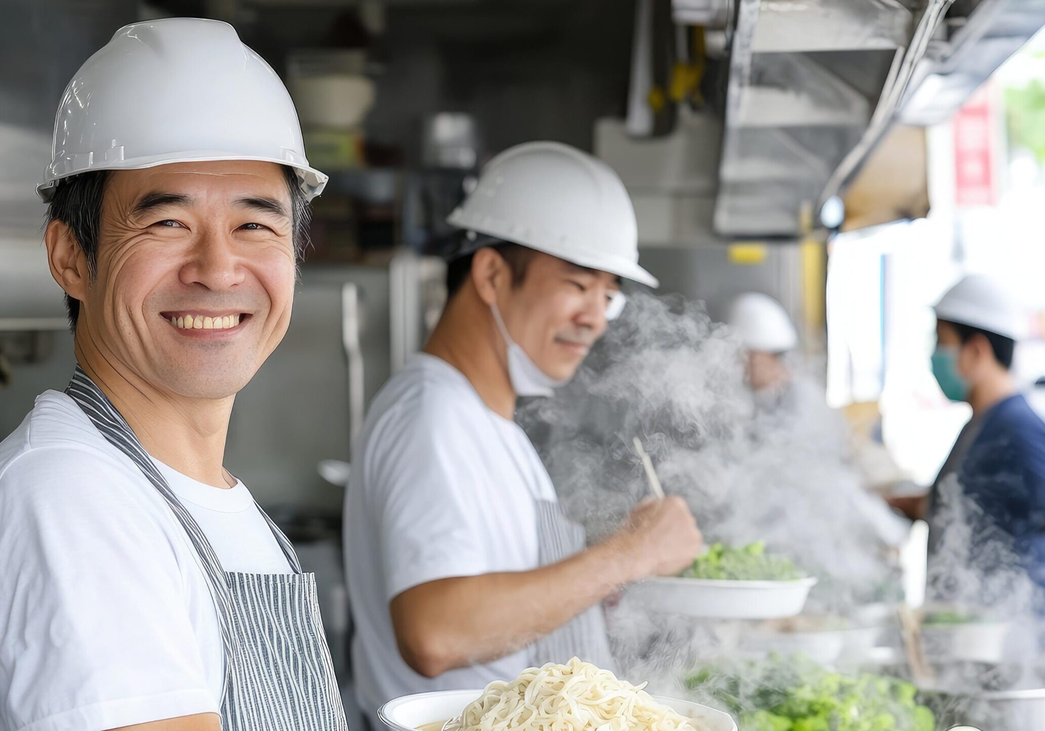 This cheerful scene features a smiling man in the foreground wearing a white hard hat and striped apron, presenting a bowl of noodles. Behind him, colleagues in matching safety gear are actively cooking amidst rising steam at a bustling outdoor food stall.