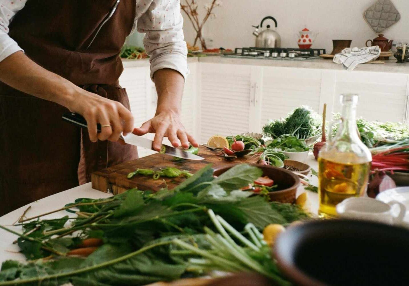In a sunlit kitchen, a person wearing a brown apron carefully slices green vegetables on a wooden cutting board. The counter is overflowing with fresh ingredients, including leafy greens, radishes, and herbs, creating a vibrant and wholesome cooking scene.