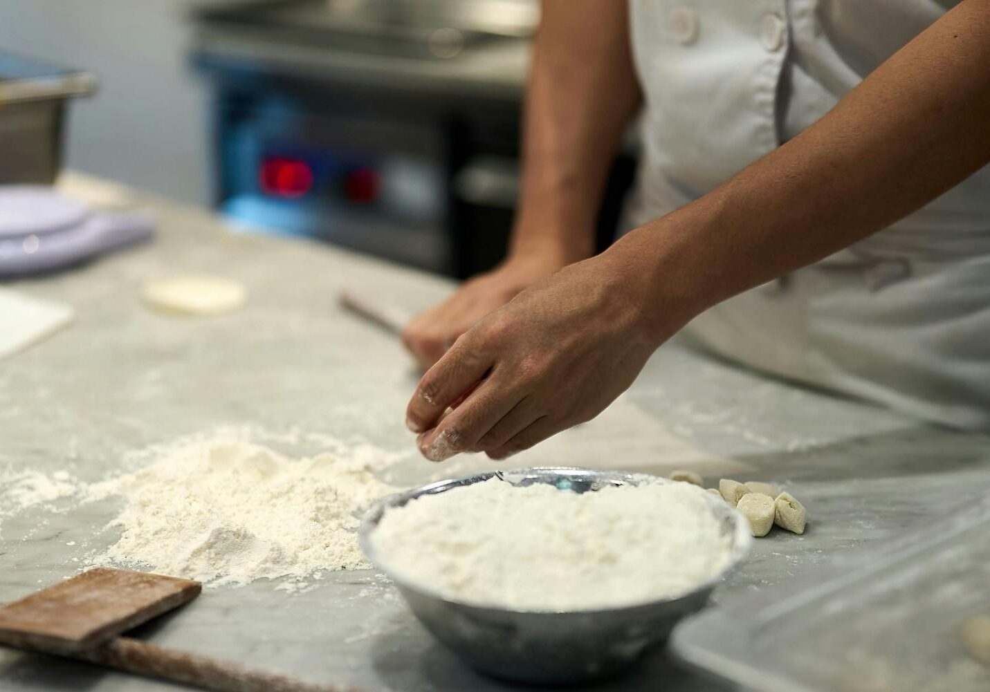A chef's hands are shown meticulously working with flour on a marble countertop, with a large bowl of white flour prominent in the foreground. Small pieces of shaped dough lie nearby, set against the blurred backdrop of a professional stainless steel kitchen.