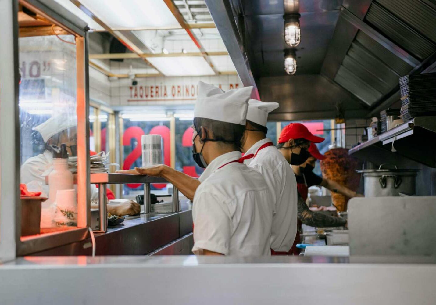 A team of kitchen staff wearing white uniforms, red aprons, and protective face masks are shown working together behind a stainless steel counter. The bustling environment appears to be a taqueria, indicated by the background signage and a traditional vertical rotisserie.