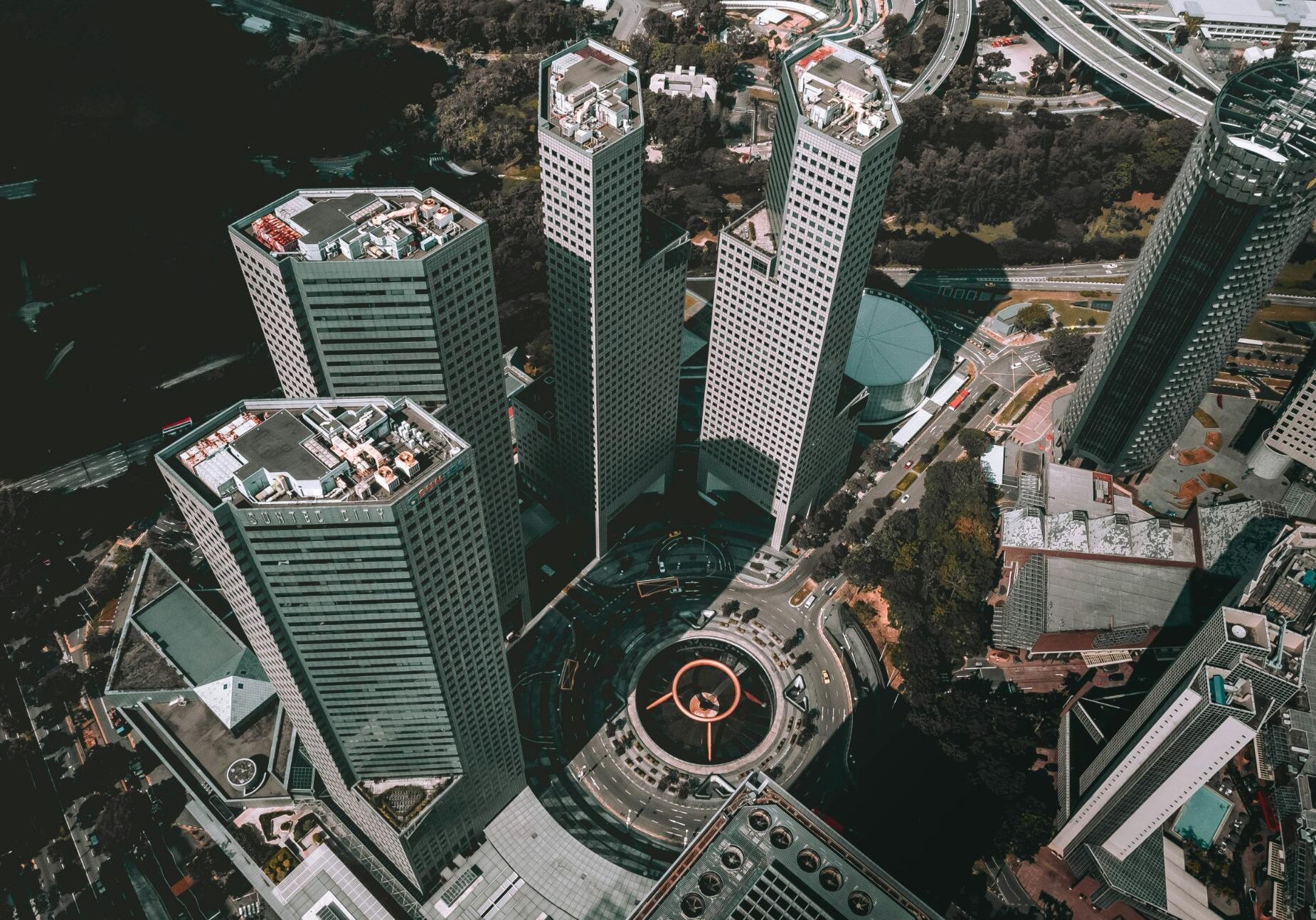 Dining Suntec - 1 This high-angle aerial photograph captures a cluster of modern skyscrapers arranged around a large, central traffic roundabout. At the heart of the circle lies a distinctive ring-shaped fountain structure, anchoring the busy urban landscape of roads and greenery.