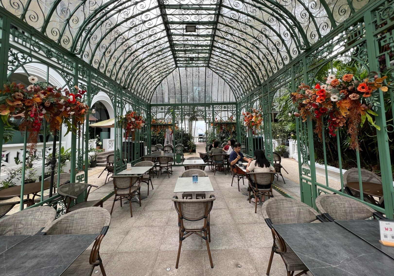 Dining Suntec - 2 This image depicts an elegant outdoor dining area enclosed by an ornate green metal trellis with an arched, scroll-patterned roof. Large autumnal floral arrangements decorate the pillars, framing the patio where patrons are seated at wicker-style chairs enjoying their meals.
