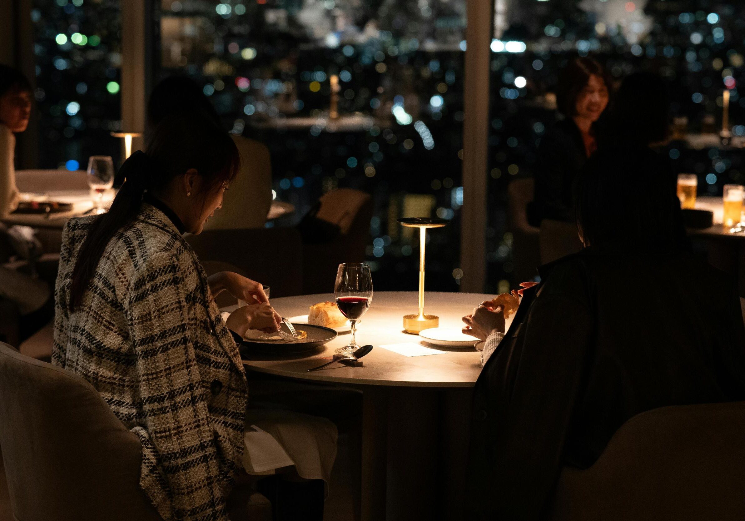 Dining Suntec - 3 Two women enjoy an intimate dinner at a dimly lit table featuring a warm lamp and glasses of red wine. Behind them, expansive windows reveal a stunning, blurred backdrop of twinkling city lights at night.