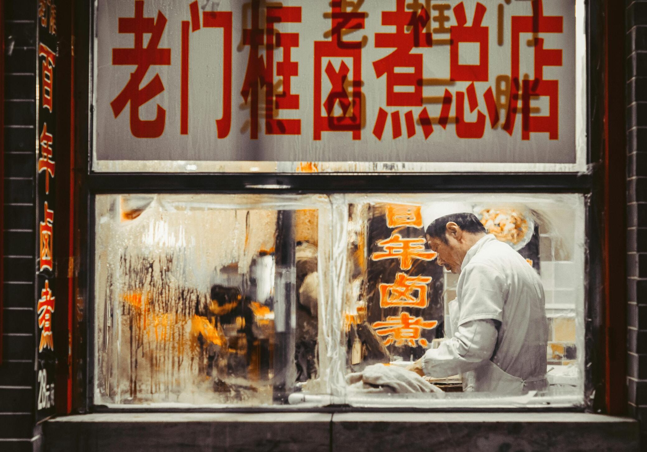 Dining Suntec - 4 Through a steamy, condensation-fogged window, a chef in a white uniform is seen busily preparing food. Large red Chinese characters dominate the storefront glass, framing the warm, bustling kitchen scene within.