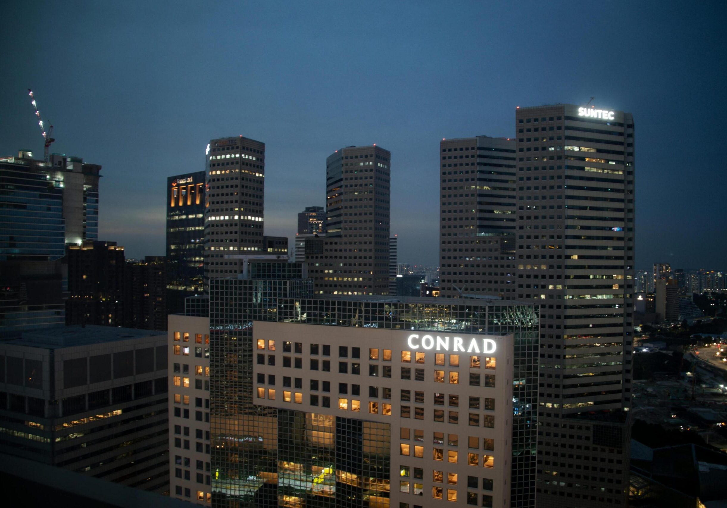 Dining Suntec - 5 This twilight cityscape captures a dense cluster of modern high-rise buildings, with their illuminated windows contrasting against the deep blue evening sky. The scene is anchored by prominent glowing signage atop the towers, specifically identifying the Conrad hotel and Suntec City, which mark this as a bustling commercial and hospitality district.
