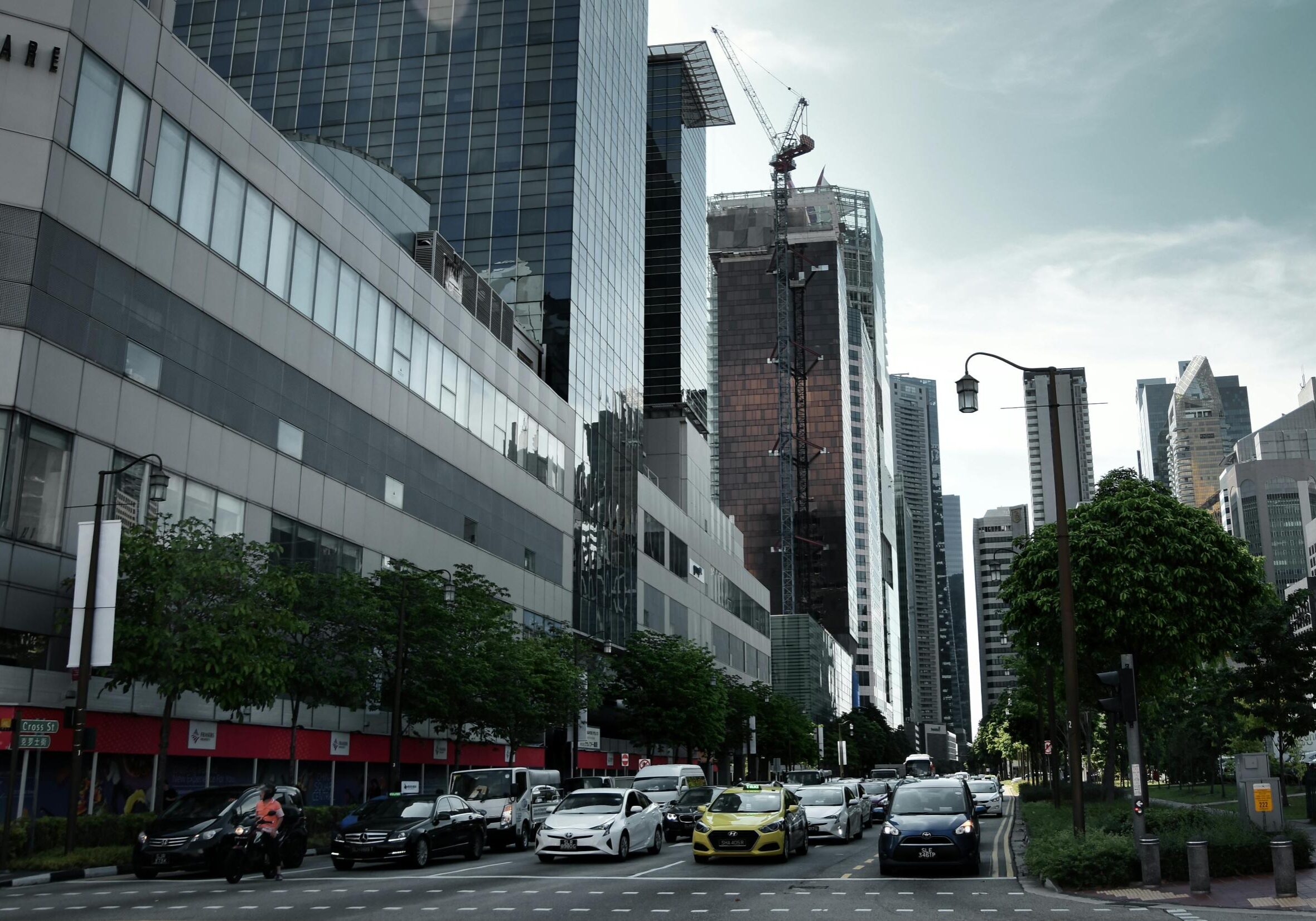 Dining Suntec - 6 This street-level view captures a busy urban intersection where a queue of cars and taxis waits beneath a skyline of towering glass office buildings. Construction cranes are visible against the overcast sky, indicating ongoing development, while rows of green trees line the road to add a touch of nature to the modern city scene.