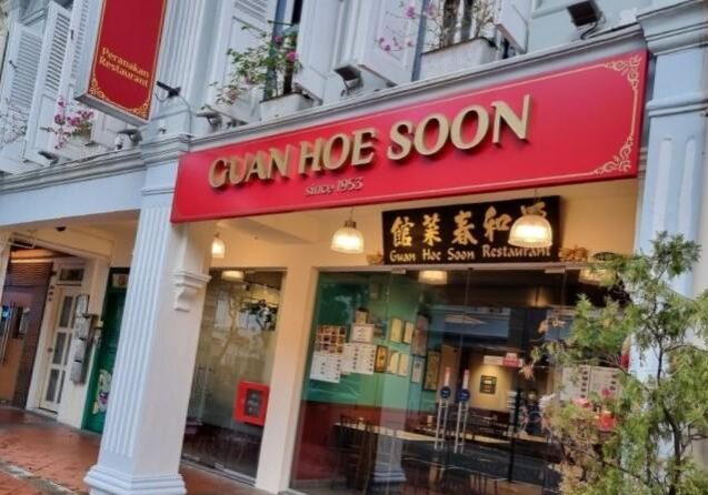 The storefront of Guan Hoe Soon Restaurant features a bold red sign with gold lettering and white fluted columns characteristic of a traditional shophouse. A secondary black sign displays the name in Chinese characters above glass doors that reveal a brightly lit, modern dining area.