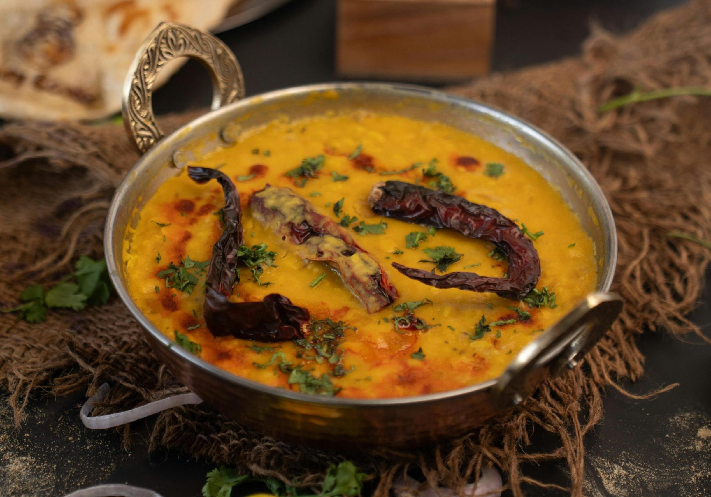 Halal Meat Restaurant Singapore - 2 This close-up image features a traditional hammered metal bowl filled with creamy yellow lentil curry, garnished with whole dried red chilies and chopped fresh green herbs. Set on a rustic burlap cloth with a hint of flatbread in the background, the presentation highlights the warm, rich textures of the dish.