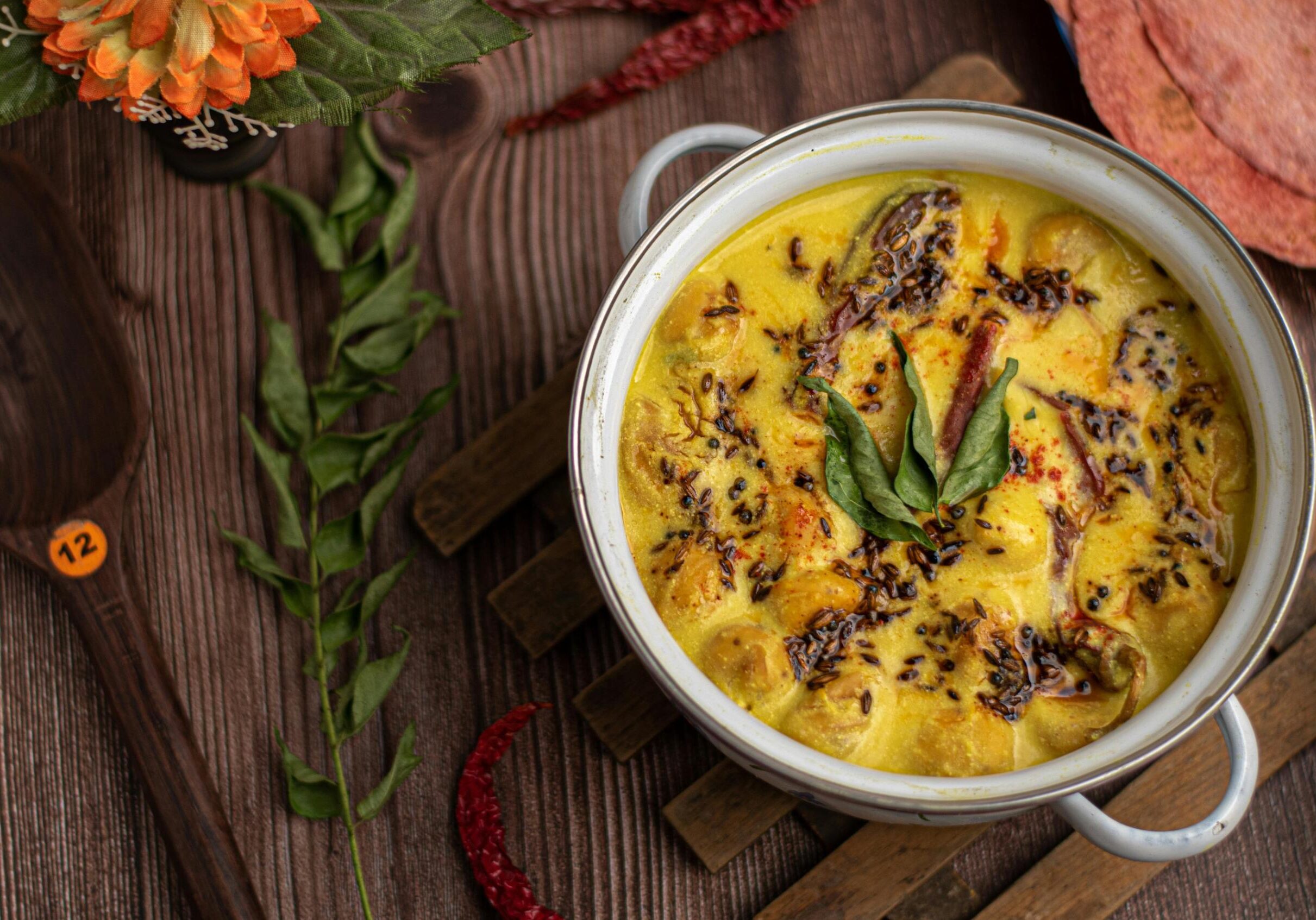 Halal Meat Restaurant Singapore - 4 This overhead shot captures a white bowl filled with creamy yellow Kadhi and dumplings, tempered with aromatic spices like cumin and dried red chilies and garnished with fresh curry leaves. Set against a dark wooden background with rustic props like a wooden spoon and pink flatbreads, the presentation is vibrant and inviting.