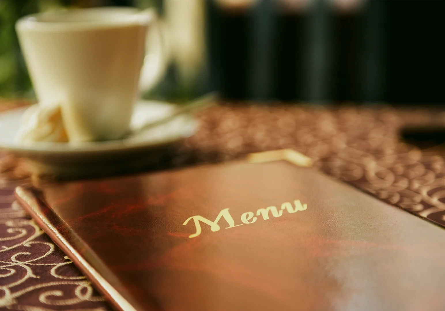 Shallow depth‑of‑field close‑up shot of a restaurant menu book on a patterned table, with the word “Menu” in focus and a cup of coffee softly blurred in the background.