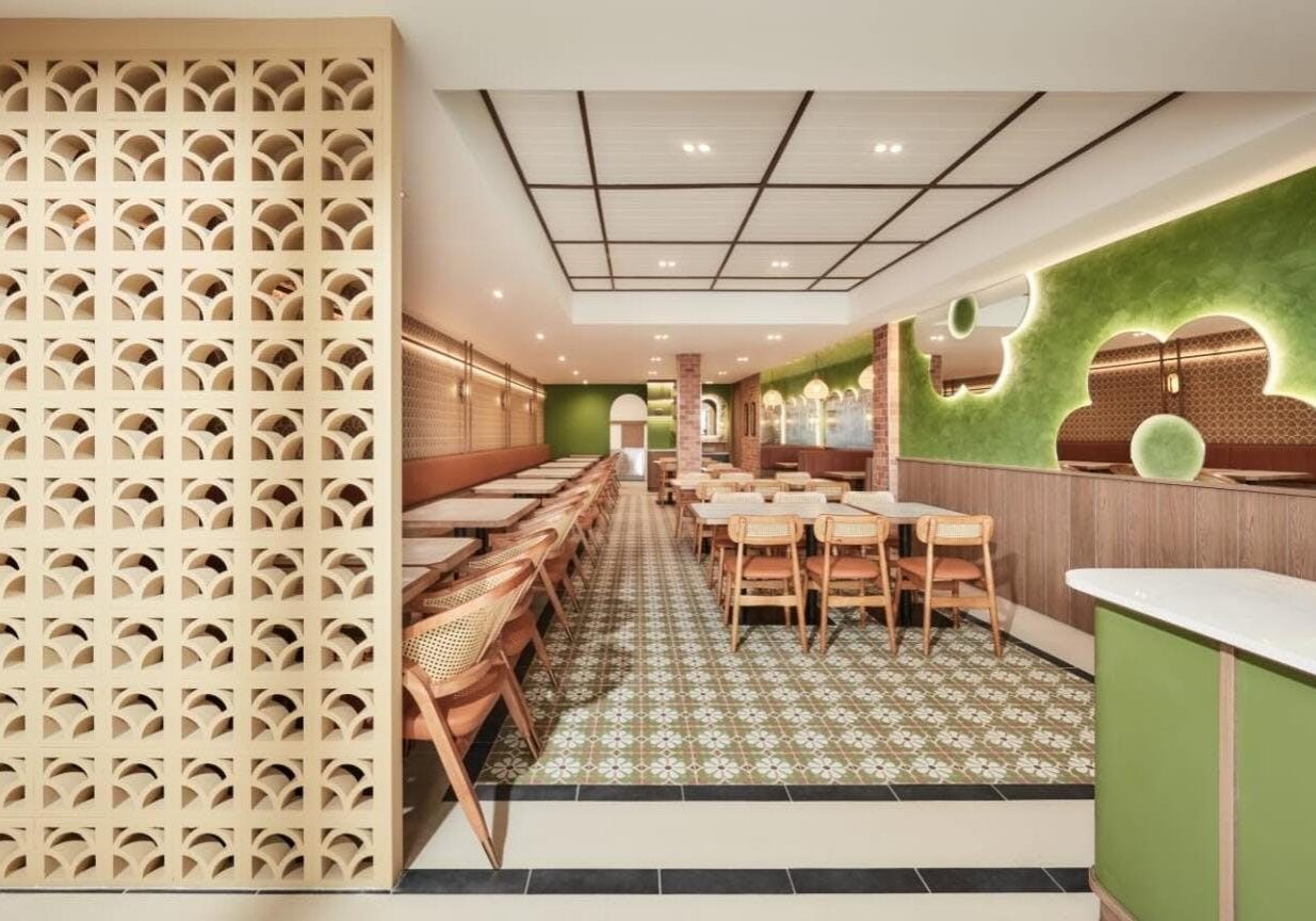 This restaurant interior features a long, narrow dining hall with floral-patterned floor tiles and a decorative breeze block partition wall on the left. The design incorporates a vibrant green accent wall with cloud-shaped mirrors and warm wood paneling, creating a bright and modern atmosphere.