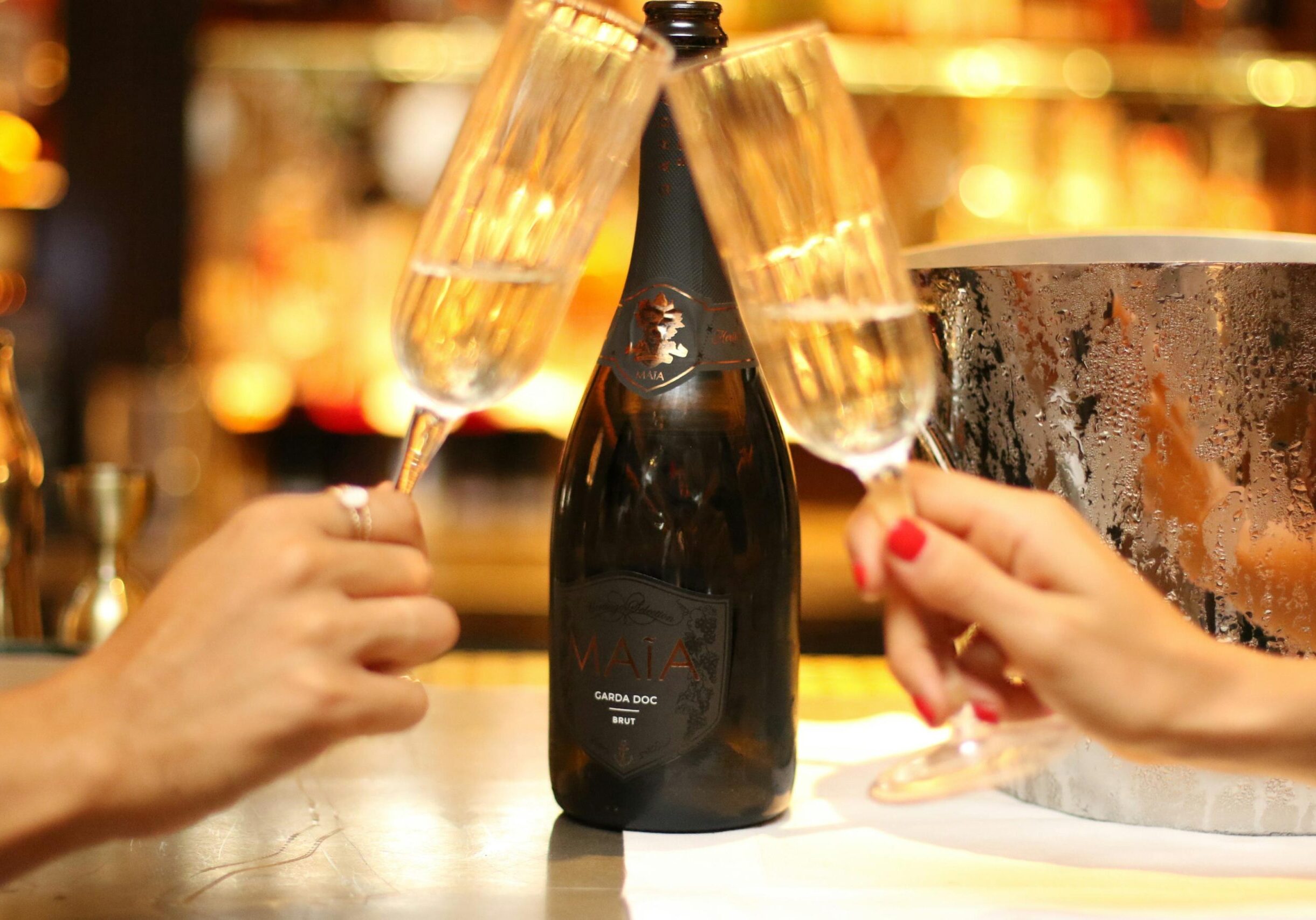 Two hands clink champagne flutes together in a celebratory toast, framing a central bottle of Maia Garda DOC Brut sparkling wine. The scene is set against a warm, bokeh-filled background with a chilled, condensation-covered ice bucket visible on the right.