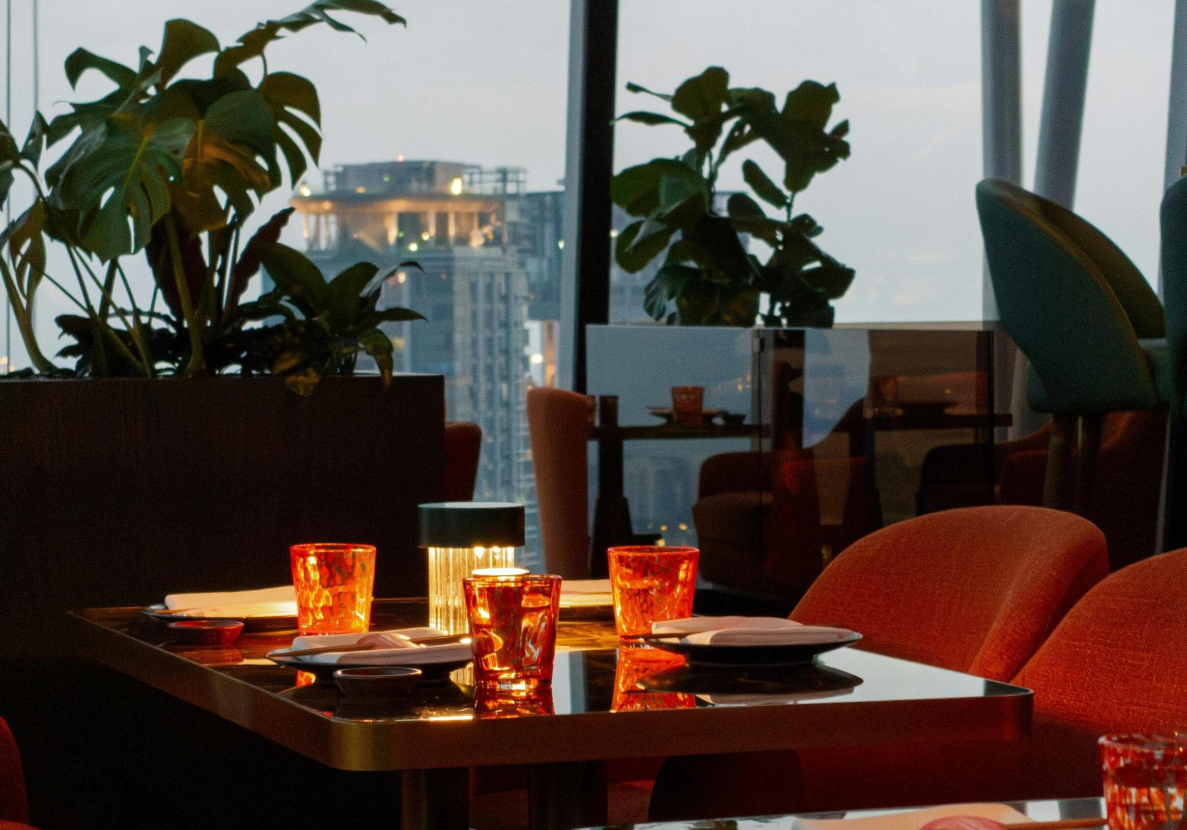 An intimate evening dining scene is set on a glossy table featuring glowing red glasses and a small lamp, surrounded by plush reddish-orange chairs. In the background, large potted plants frame floor-to-ceiling windows that offer a view of a city skyline at dusk.