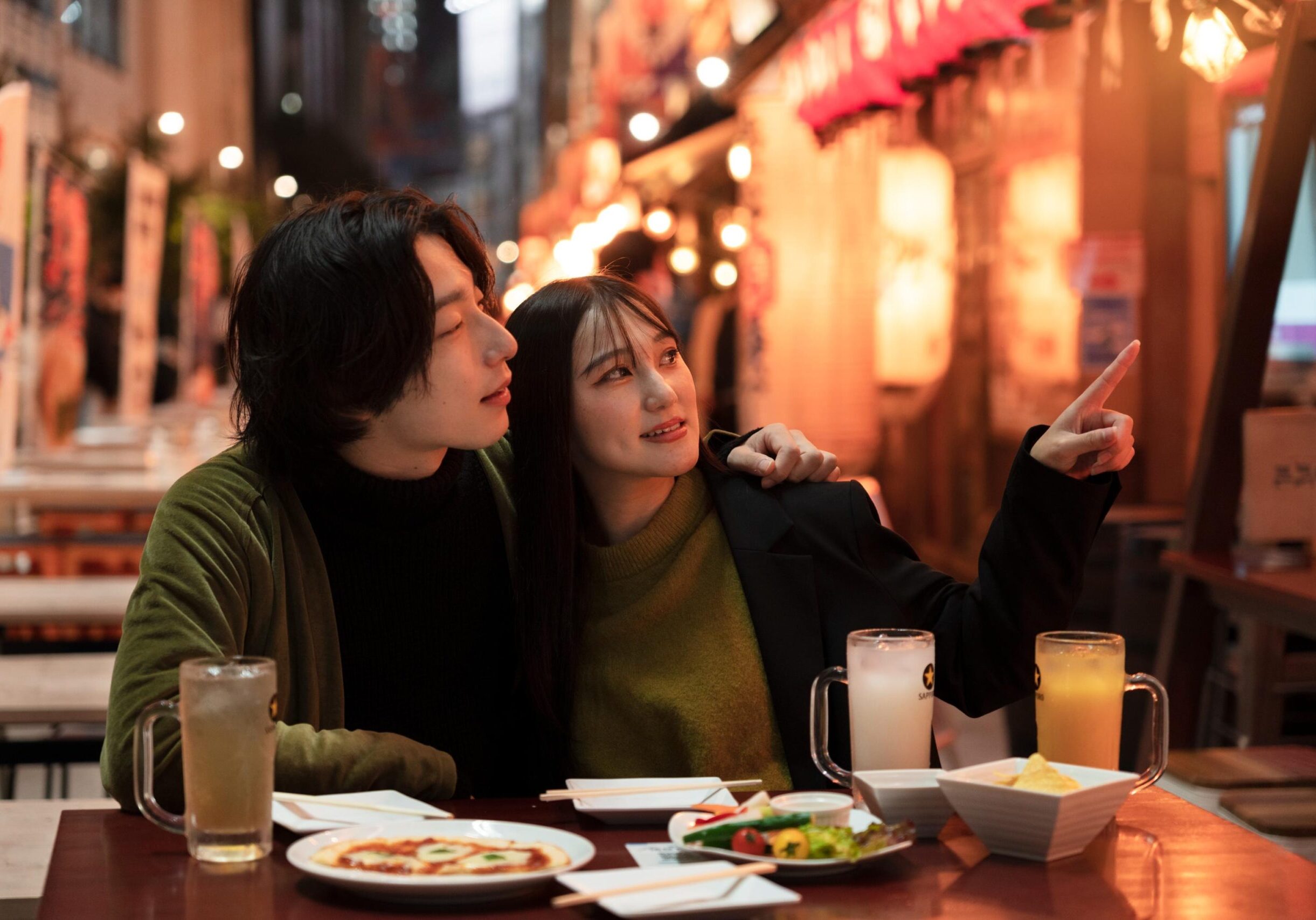 A young couple sits closely together at an outdoor table, with the woman pointing toward something in the distance while her companion looks on attentively. The vibrant night scene is illuminated by the warm glow of hanging lanterns, surrounding their table filled with large mugs of drinks and plates of food.