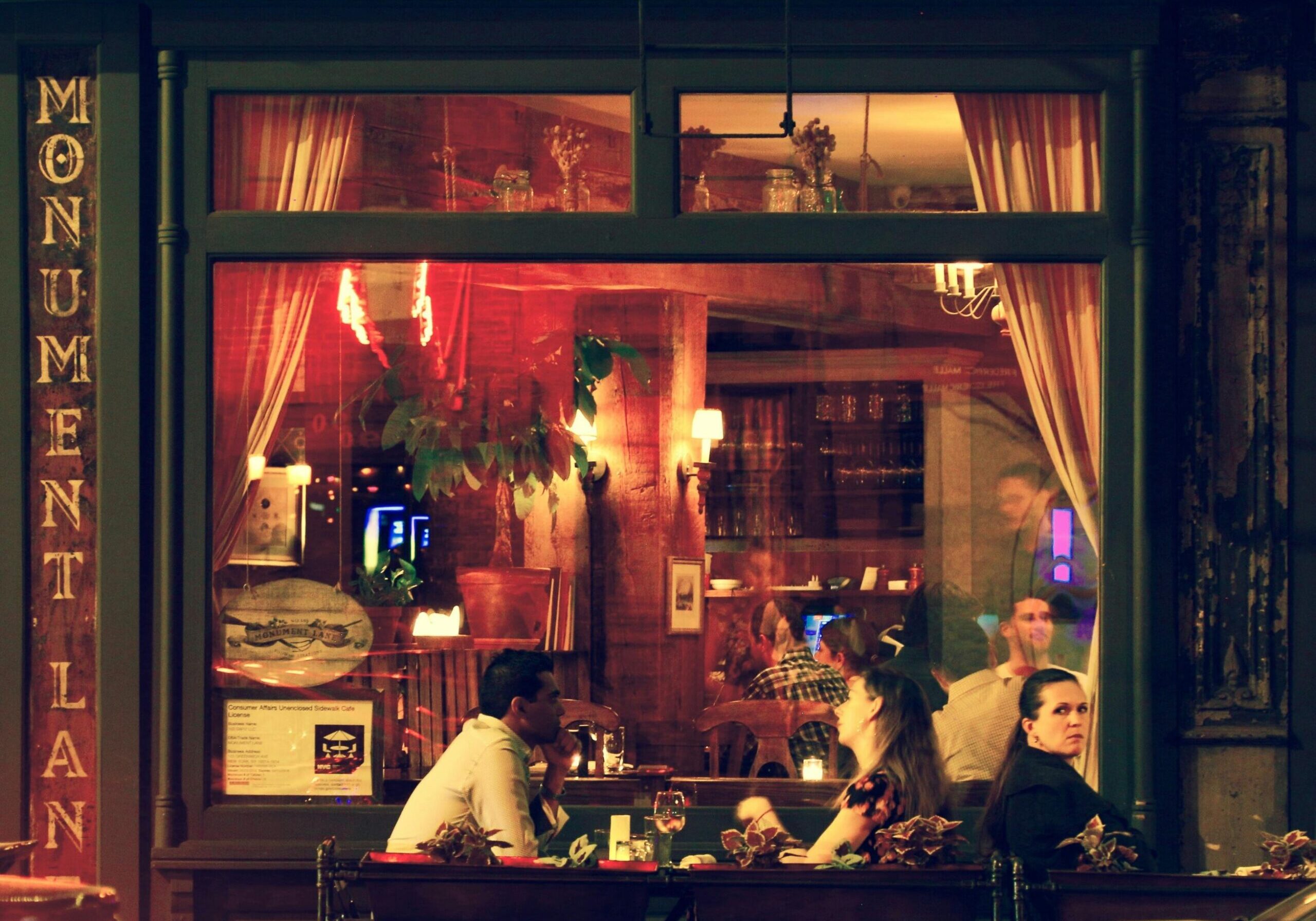 A vertical sign reading "Monument Lane" frames the left side of a bustling restaurant storefront, viewed through a large glass window. Inside, diners are engaged in conversation under warm lighting, surrounded by hanging plants and rustic decor that create a cozy evening atmosphere.