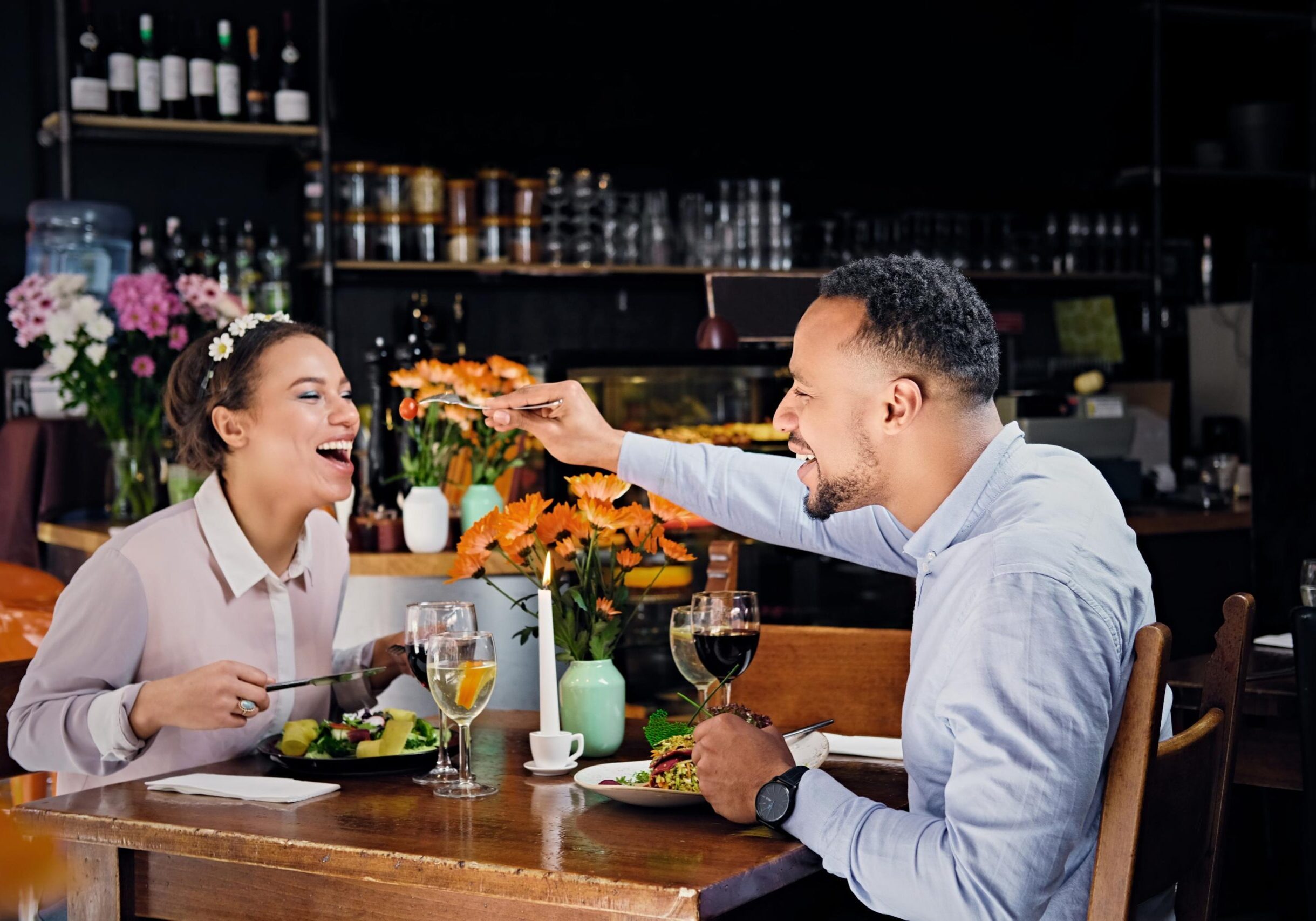 A joyful couple enjoys a meal at a rustic wooden table, where a smiling man playfully feeds a cherry tomato to his laughing companion. The table is adorned with plates of fresh salad, wine glasses, and a vase of orange flowers, creating a warm and lively restaurant atmosphere.