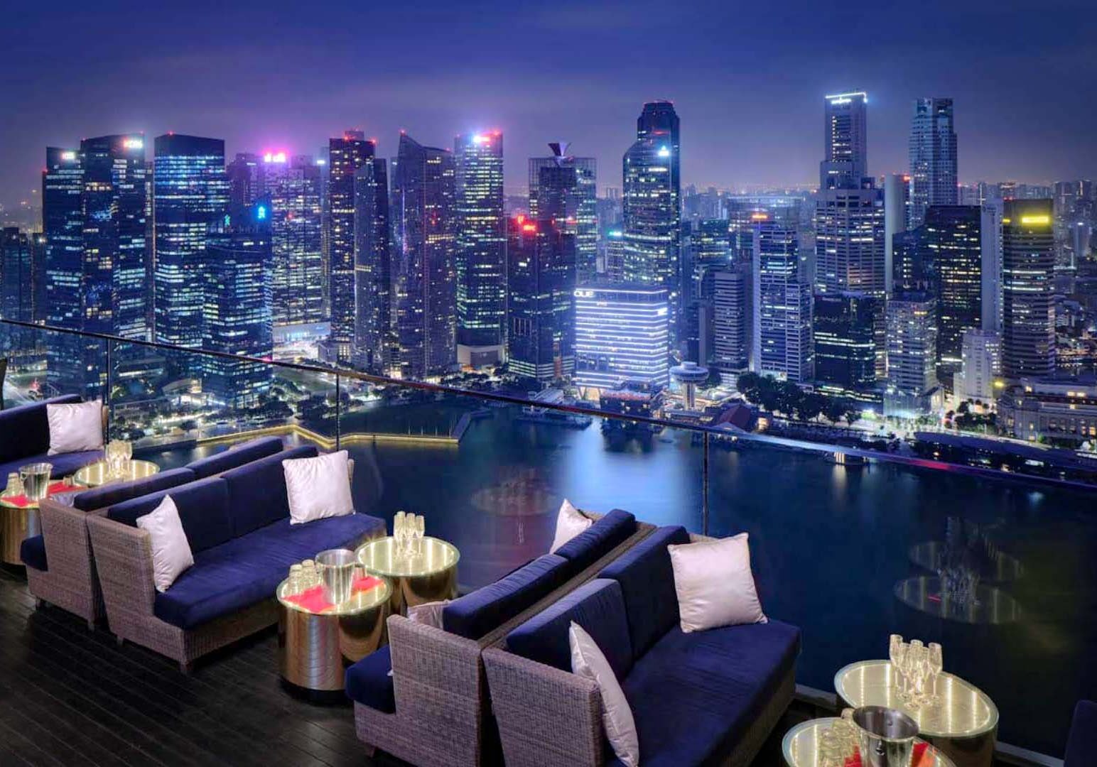 A rooftop lounge with cushioned seating overlooking a brightly lit city skyline at night, with reflections shimmering on the water below.