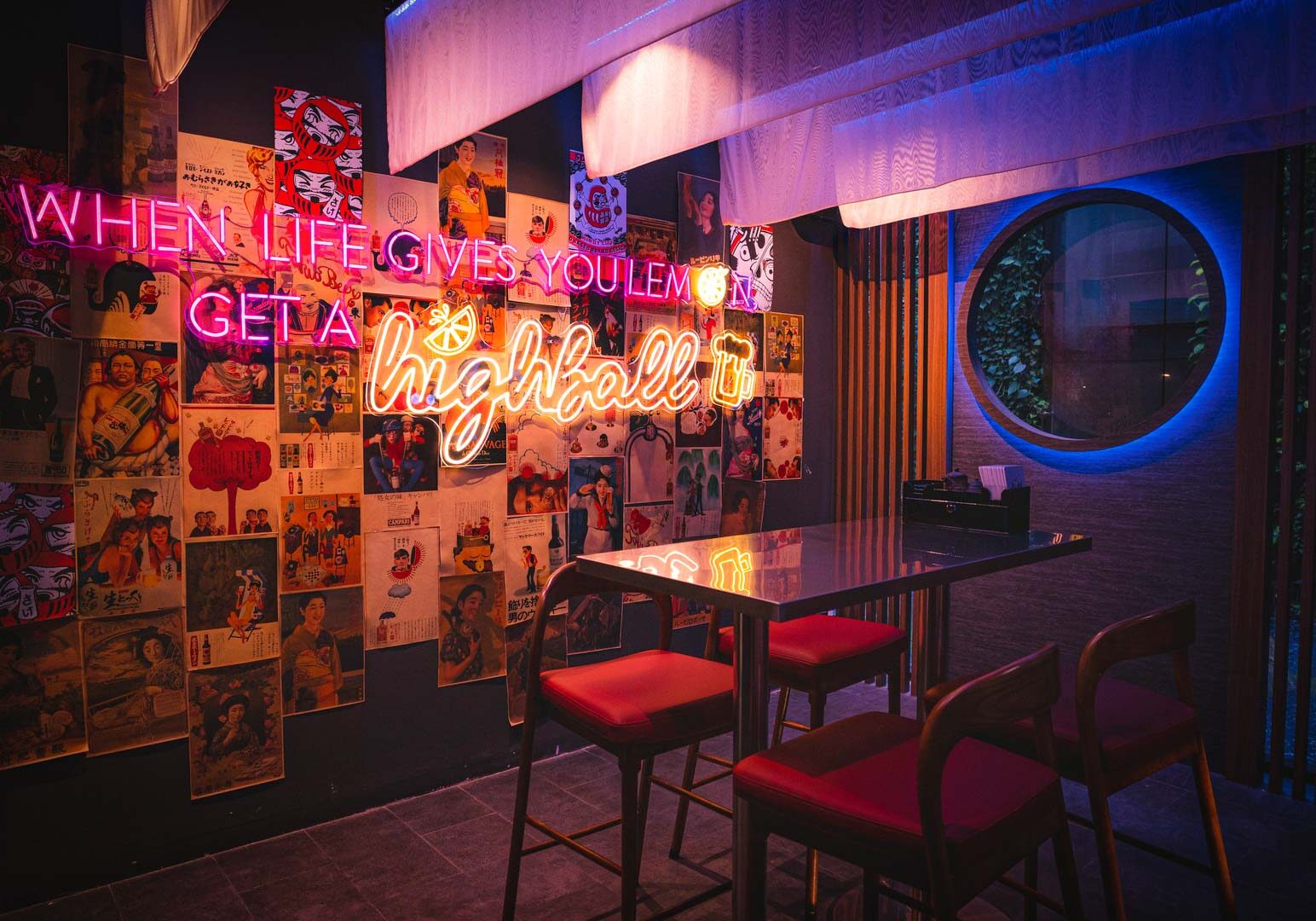 A dimly lit bar area with neon signage reading ‘When life gives you lemons, get a highball,’ displayed above a wall collage of posters, with a high table and red cushioned chairs in front.