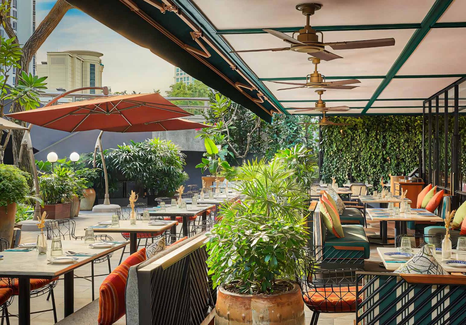 An outdoor restaurant patio surrounded by lush greenery, featuring neatly arranged tables, overhead fans, and colorful cushions under a partially covered canopy.