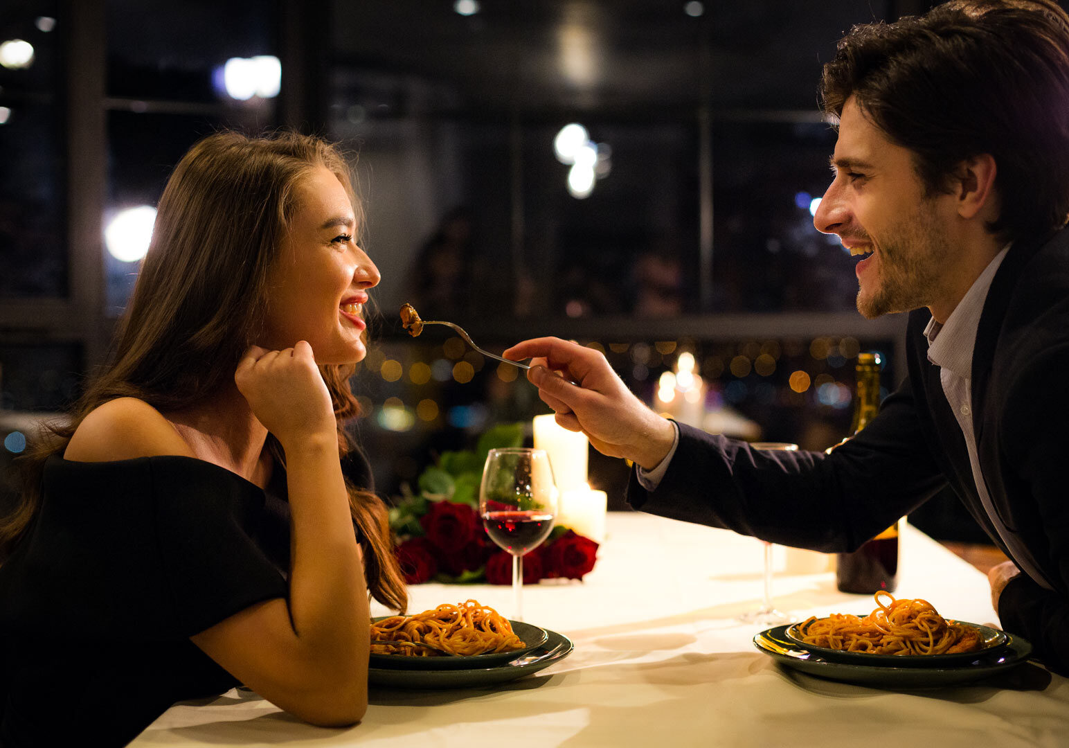 A candlelit dinner scene where one person extends a fork with a bite of pasta toward the other across a table set with plates of spaghetti, a glass of red wine, and a bouquet of roses, with city lights visible through the window in the background.
