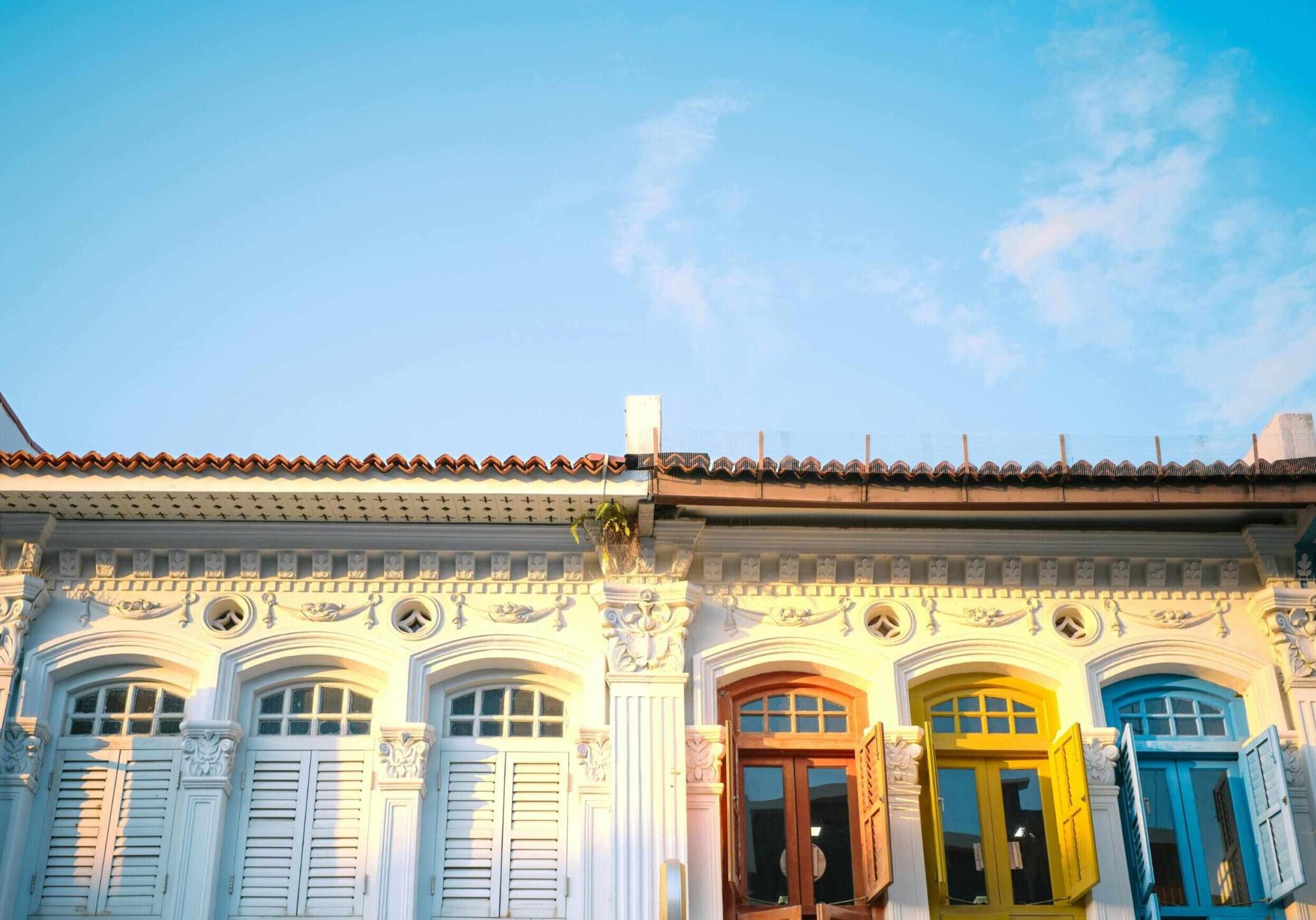 This image captures the colorful and ornate facades of traditional shophouses, likely in a Southeast Asian city like Singapore or Malaysia, under a bright blue sky.