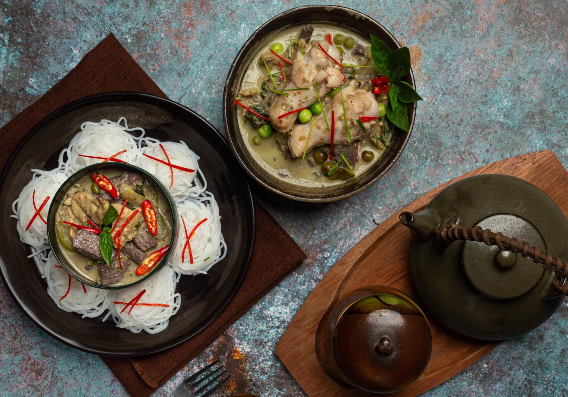 This is an overhead, dark-toned image featuring two servings of Thai Green Curry Gaeng Keow Wan presented alongside mounds of thin rice vermicelli noodles.