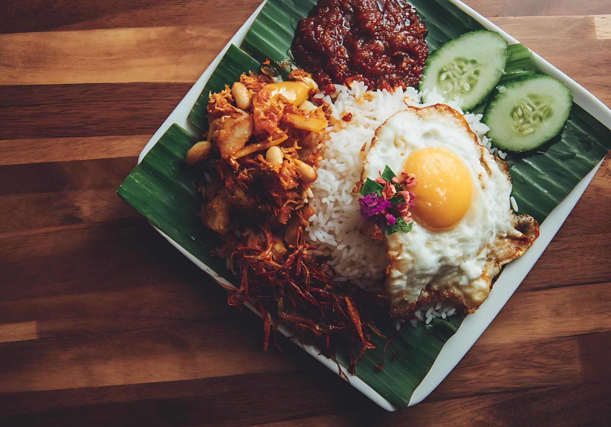 This is a close-up, top-down shot of a classic Southeast Asian rice dish, likely Nasi Lemak, served on a square white plate lined with a banana leaf and placed on a wooden table.