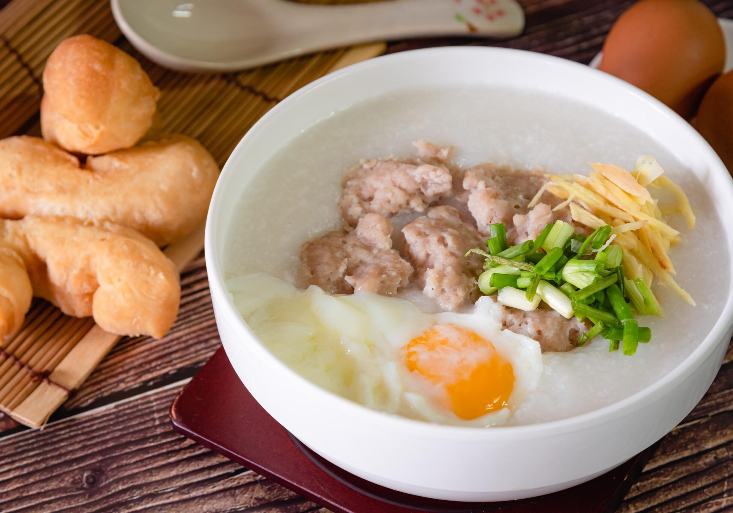 Oldest Teochew - 1 This image features a comforting white bowl of rice porridge, or congee, generously topped with seasoned pork chunks, a soft-poached egg, julienned ginger, and fresh green onions. To the left, a serving of golden-brown fried dough sticks rests on a bamboo mat, completing this classic and savory Asian breakfast arrangement.