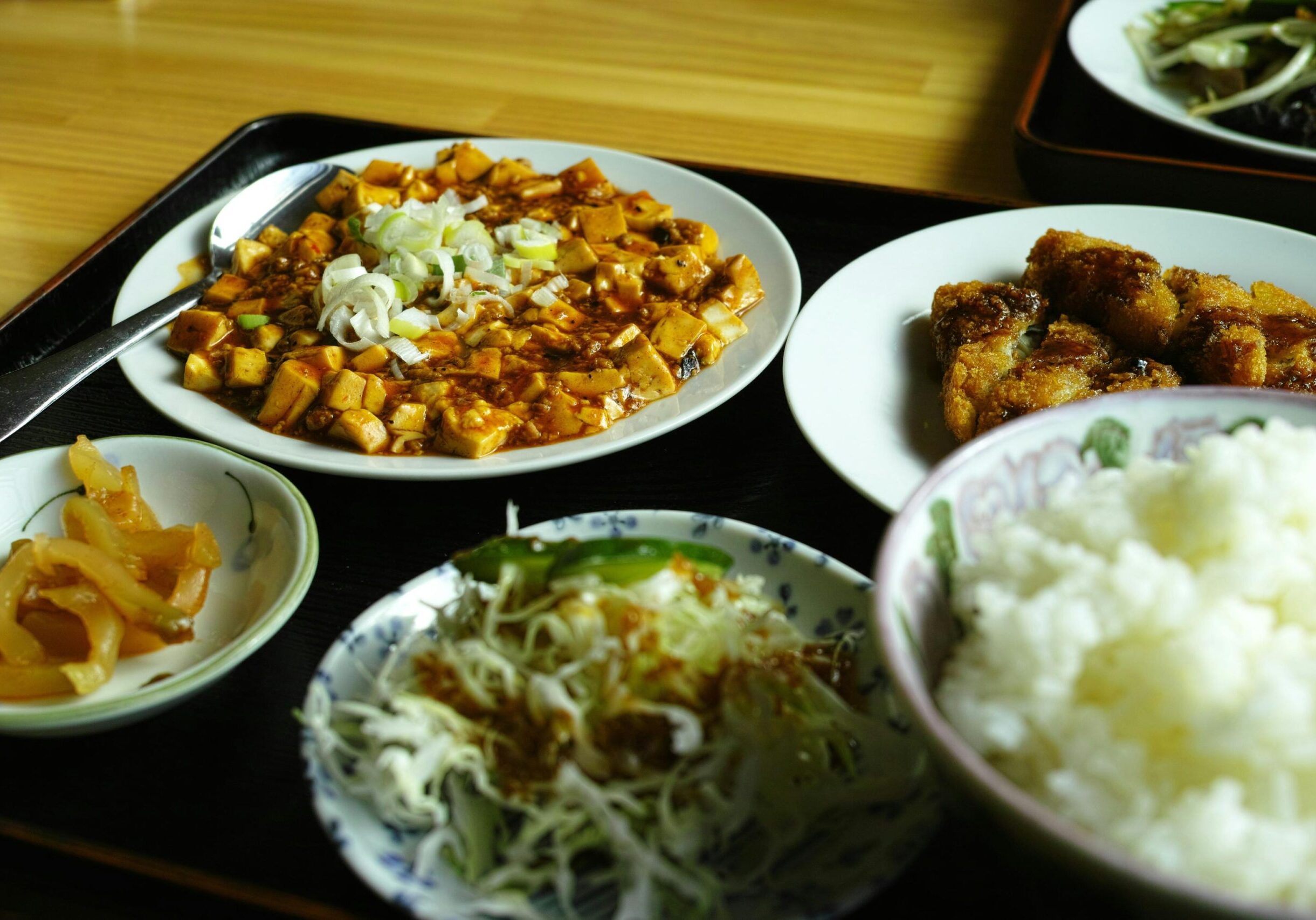 Oldest Teochew - 2 This appetizing image captures a hearty meal set centered around a plate of spicy mapo tofu topped with fresh scallions and a side of sauce-glazed fried meat. The tray is filled out with essential accompaniments, including a large bowl of fluffy white rice, a small shredded cabbage salad, and a dish of pickled vegetables.