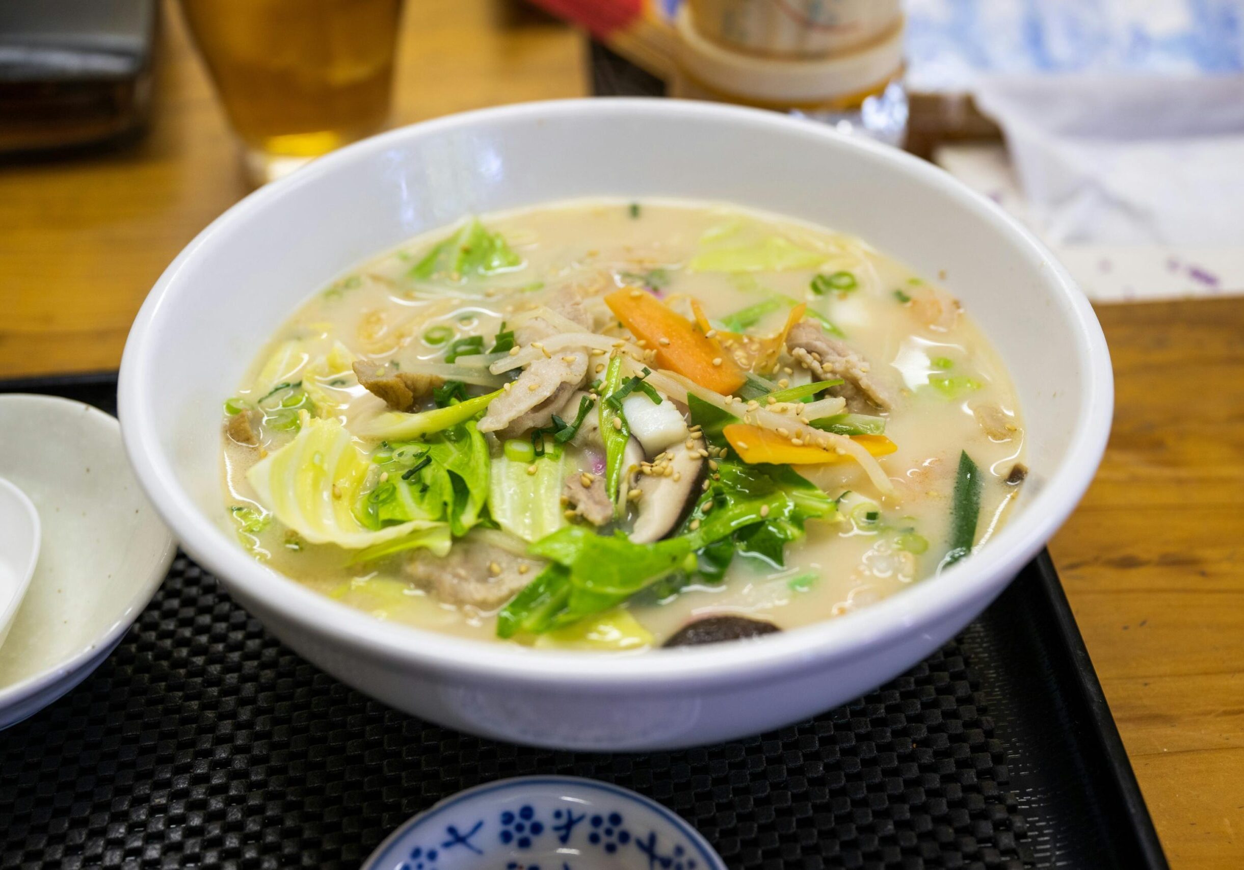 Oldest Teochew - 4 This image highlights a savory bowl of creamy teochew porridge, packed with generous toppings of cabbage, carrots, sliced pork, mushrooms, and green onions. Served on a black textured tray on a wooden table, the dish is finished with a sprinkle of sesame seeds and sits next to a white ceramic spoon.