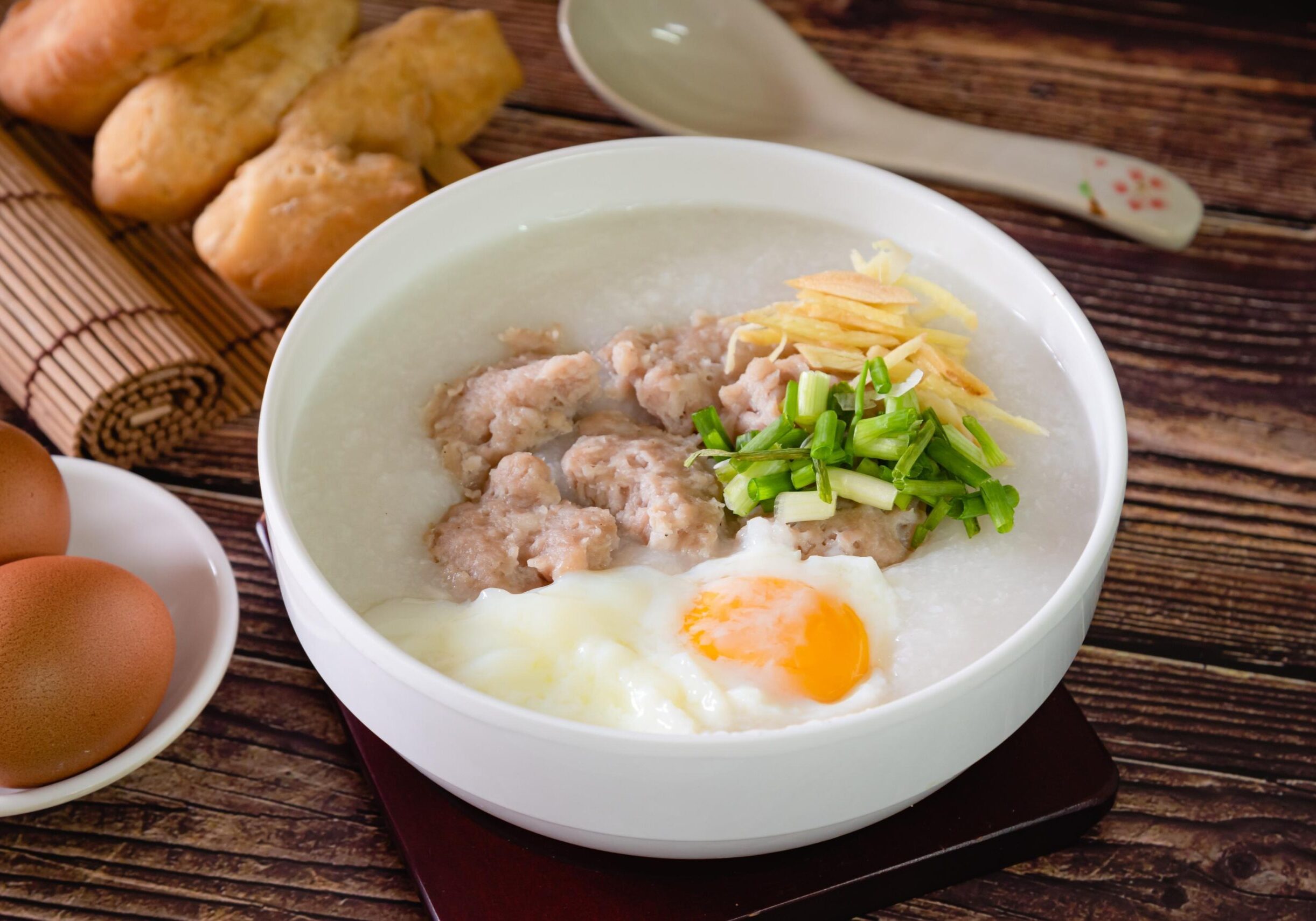 Oldest Teochew - 5 This image displays a warm bowl of white rice porridge, or congee, topped with savory pork meatballs, a soft poached egg, shredded ginger, and chopped scallions. The meal is presented on a dark wooden table alongside traditional accompaniments, including fried dough sticks resting on a bamboo mat and a small plate of whole brown eggs.