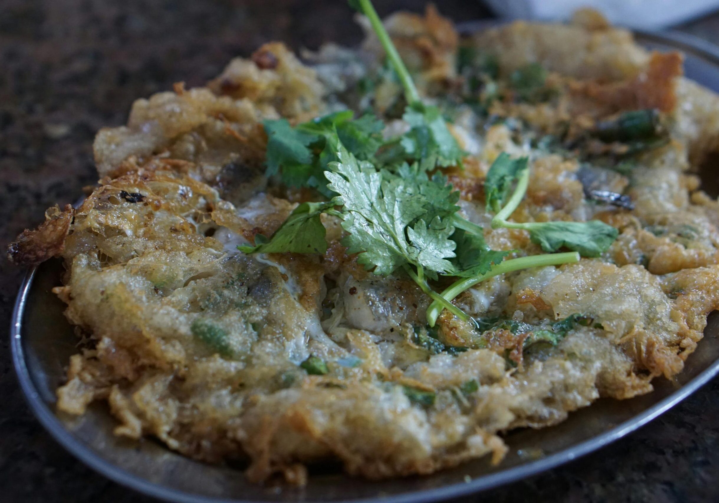 Oldest Teochew - 6 This image features a golden-brown, pan-fried omelet dish, likely an oyster omelet, characterized by its crispy edges and rich, savory texture. It is presented on a metallic oval plate and topped with a garnish of fresh green coriander sprigs to add brightness to the oily dish.